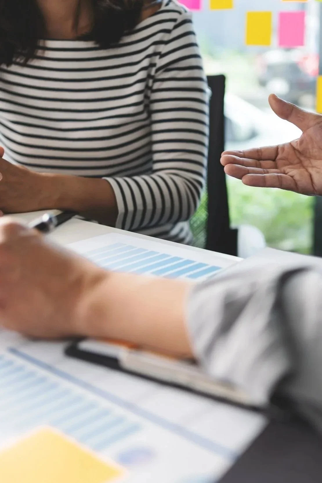 Two people are engaged in a discussion at a desk with documents and a pen. One person is wearing a black and white striped shirt, and the other’s hand is gesturing toward the documents, hinting at a collaborative or professional setting.