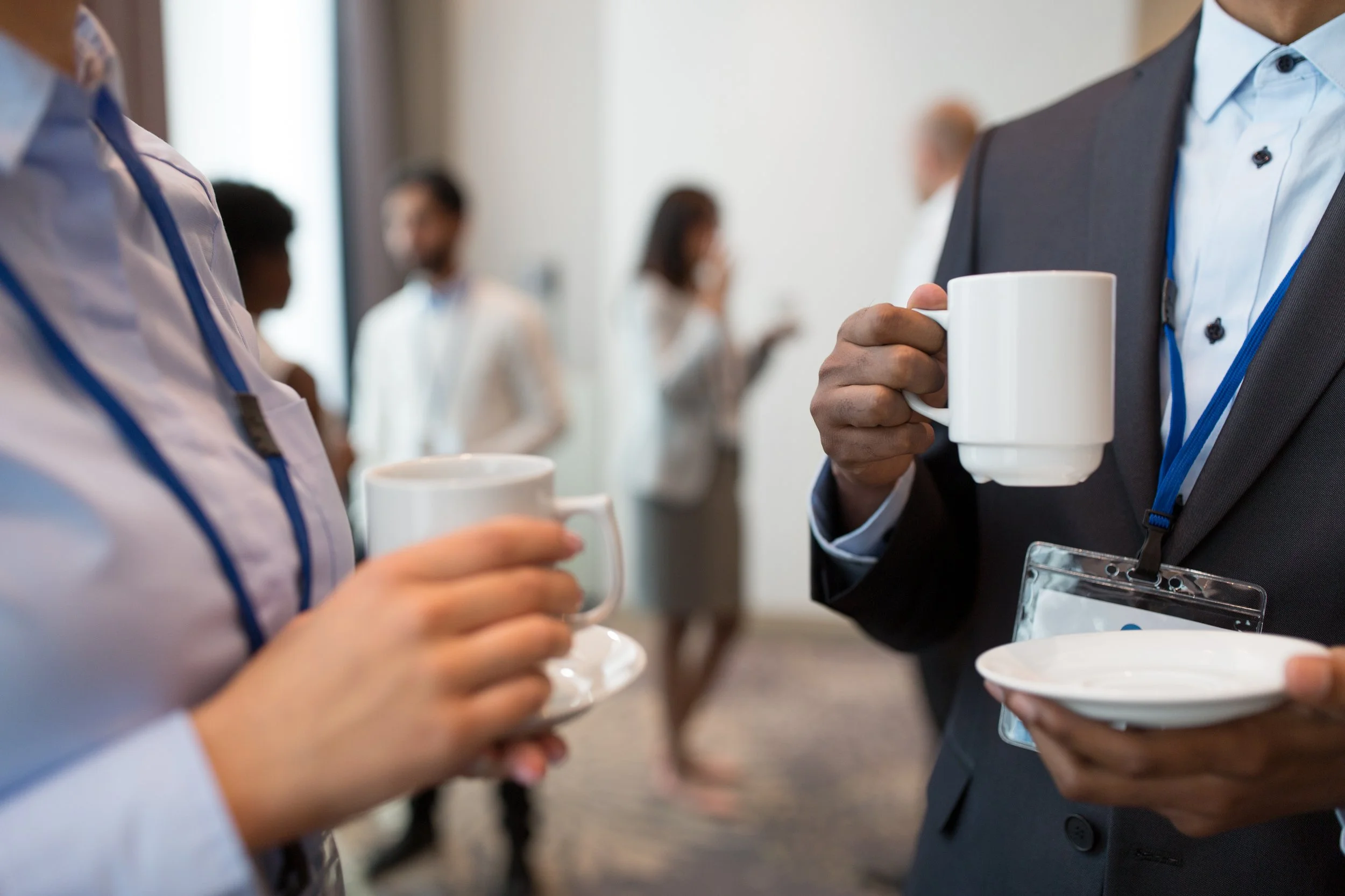 Two people in business attire sharing a coffee during a conversation at a professional event, with blurred people in the background.