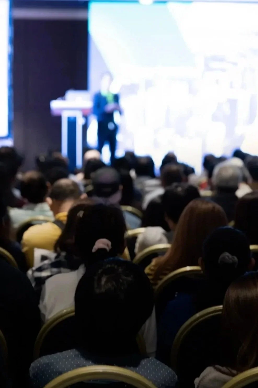 A speaker giving a presentation on a stage in front of a large audience seated in chairs.