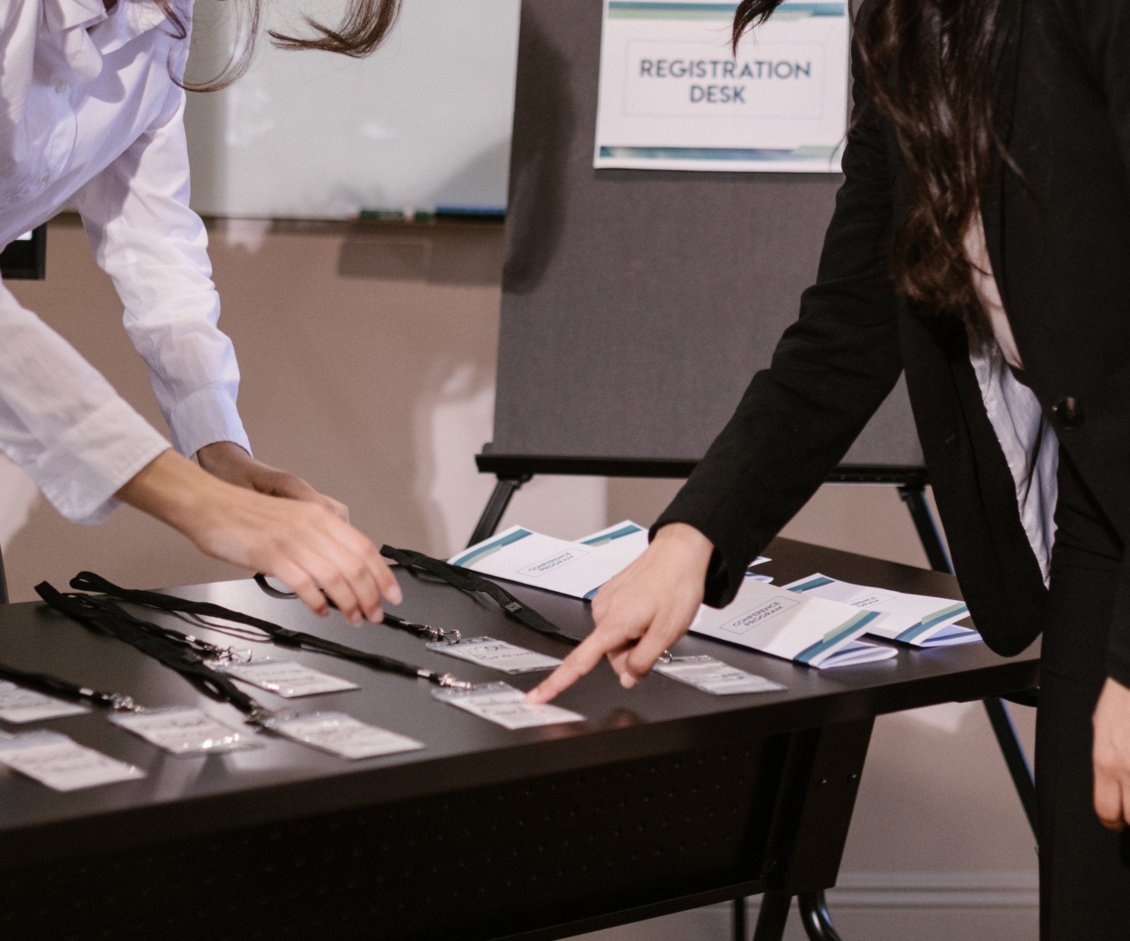 Two women at a registration desk are handling event badges or lanyards, with registration materials spread out on the table. A sign in the background reads 'Registration Desk.'