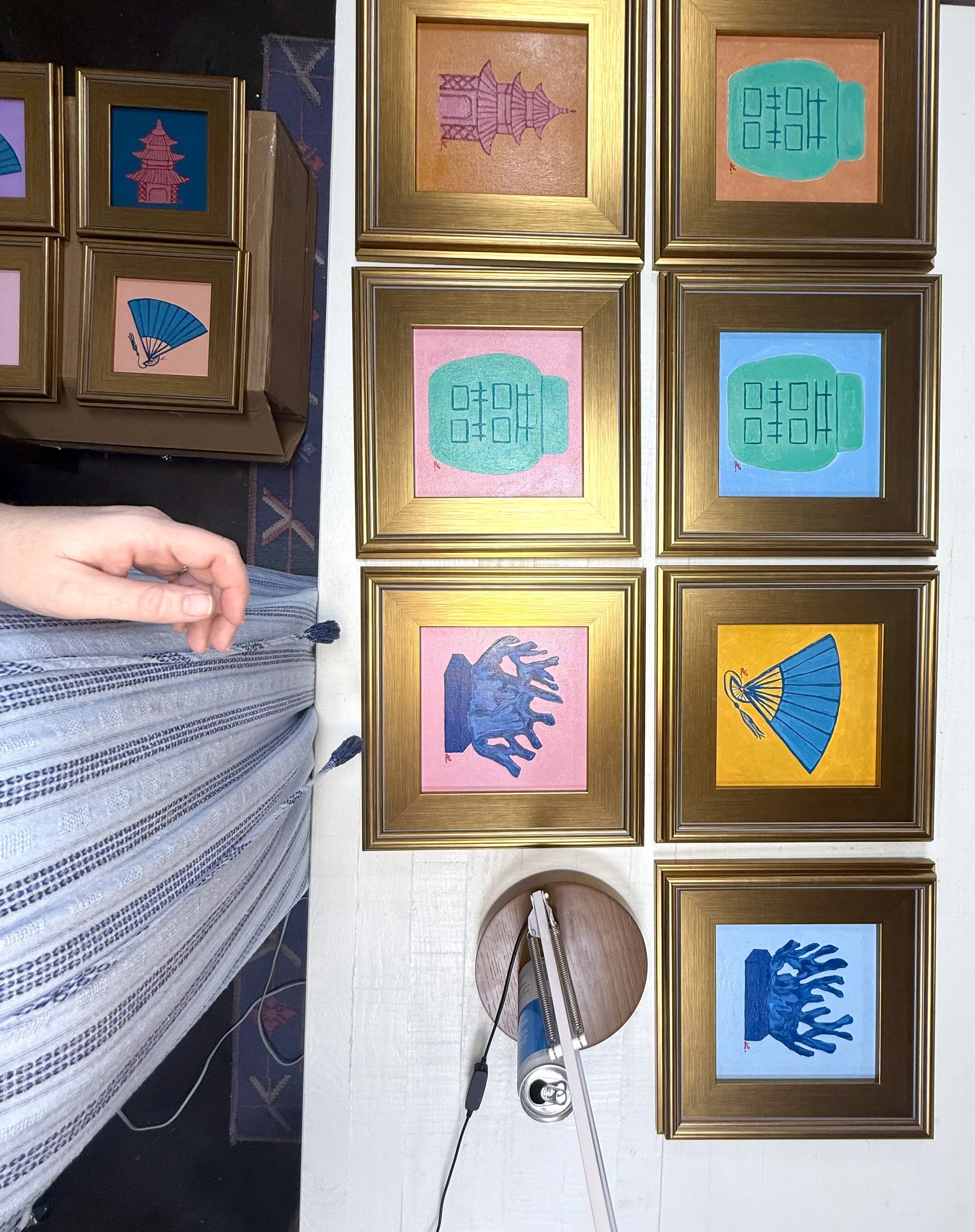 Multiple framed pictures with abstract and Asian-inspired designs on a wall, with embroidery tools and a person's hand visible in the foreground.