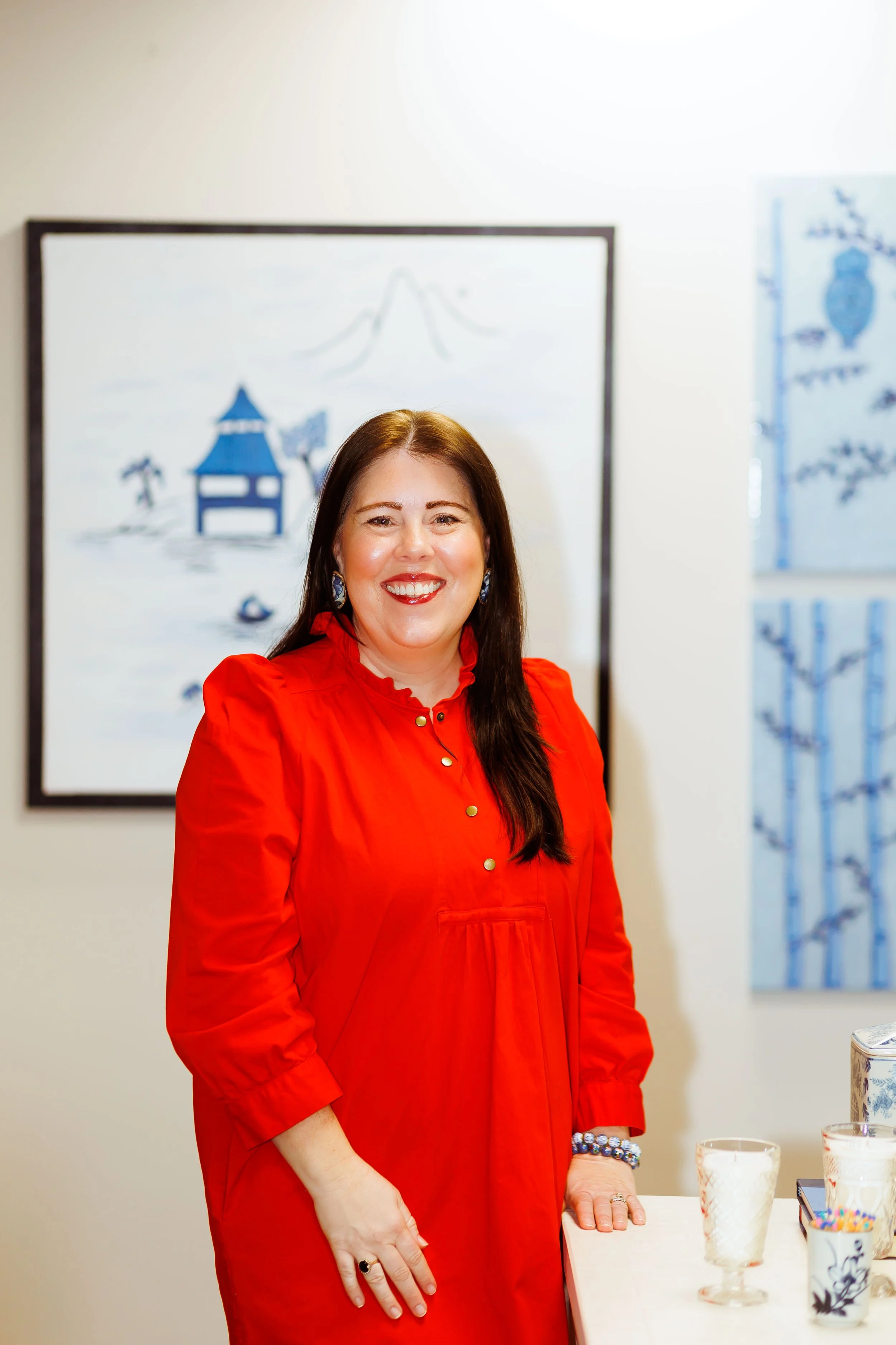 Smiling woman with long dark hair wearing a red dress standing next to a table with glasses of white beverage and a cup with colorful sprinkles, in an art gallery or exhibition space.