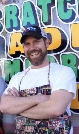 Smiling man wearing a Star Wars apron and a blue cap, standing in front of a colorful mural with large letters.