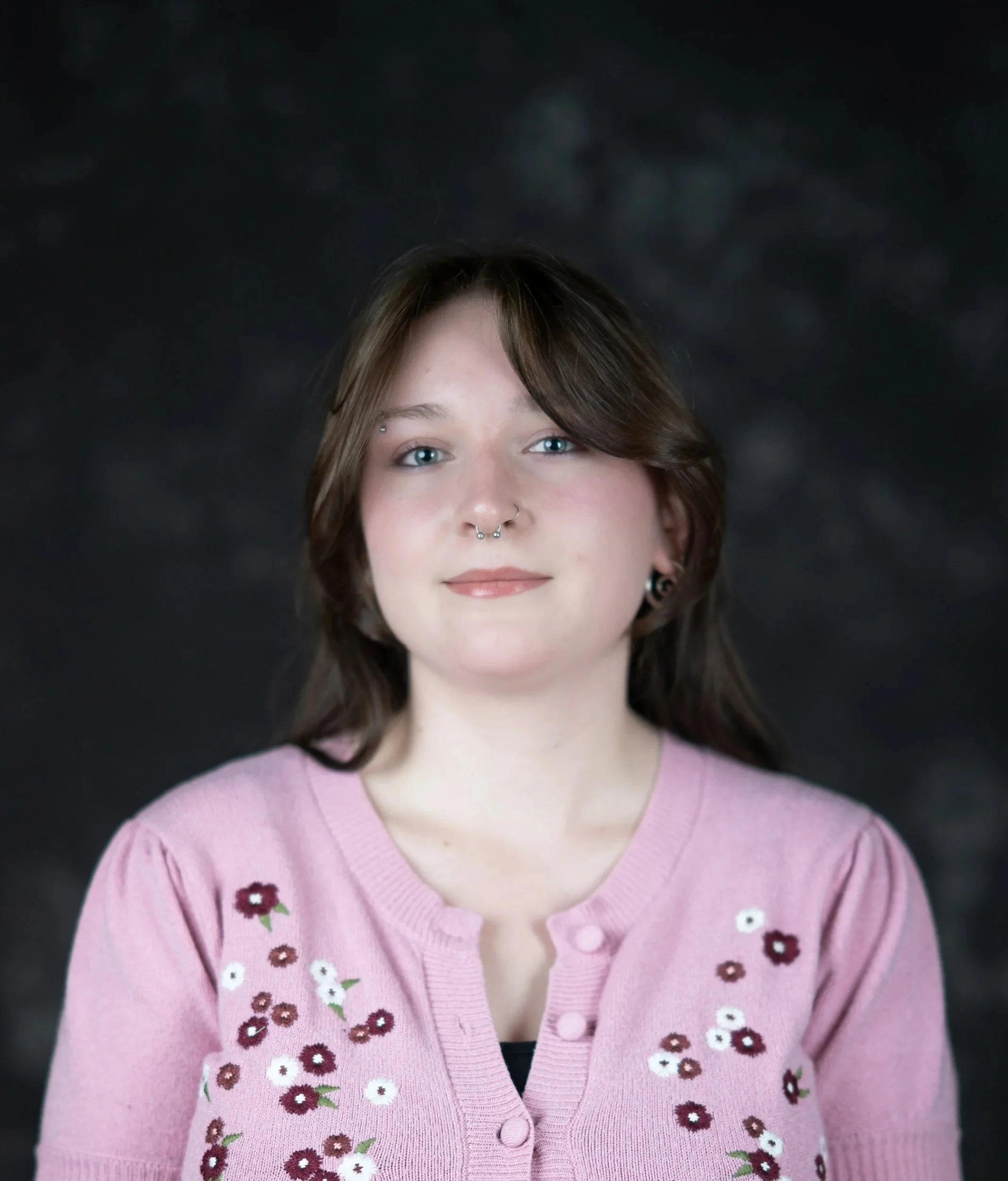 Close-up of a woman with blue eyes, short brown hair, and a septum piercing, wearing a pink cardigan with embroidered flowers against a dark background.