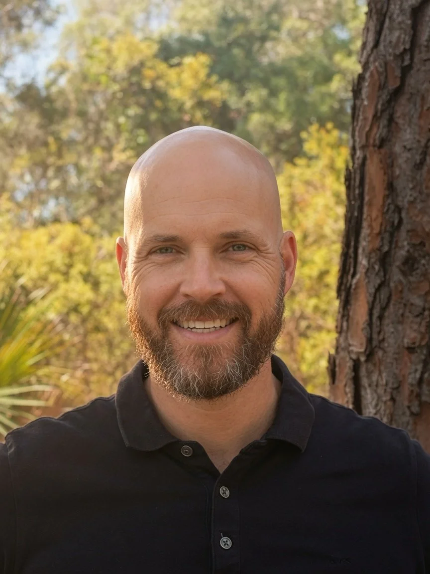 A smiling bald man with a beard wearing a black polo shirt outdoors near a tree, with blurred autumn foliage in the background.