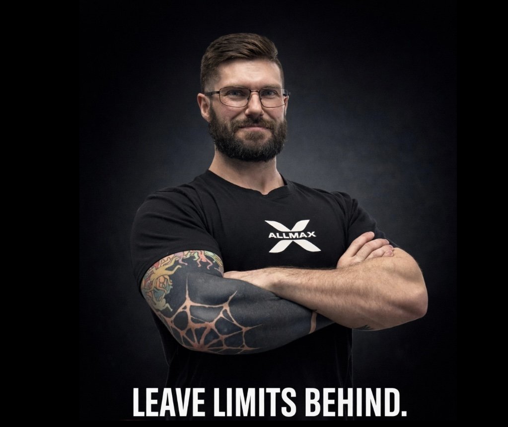 A man with glasses and a beard wearing a black t-shirt with the Allmax logo, crossing his arms, displaying a colorful tattoo sleeve on his left arm, standing against a dark background with the text 'Leave Limits Behind' at the bottom.