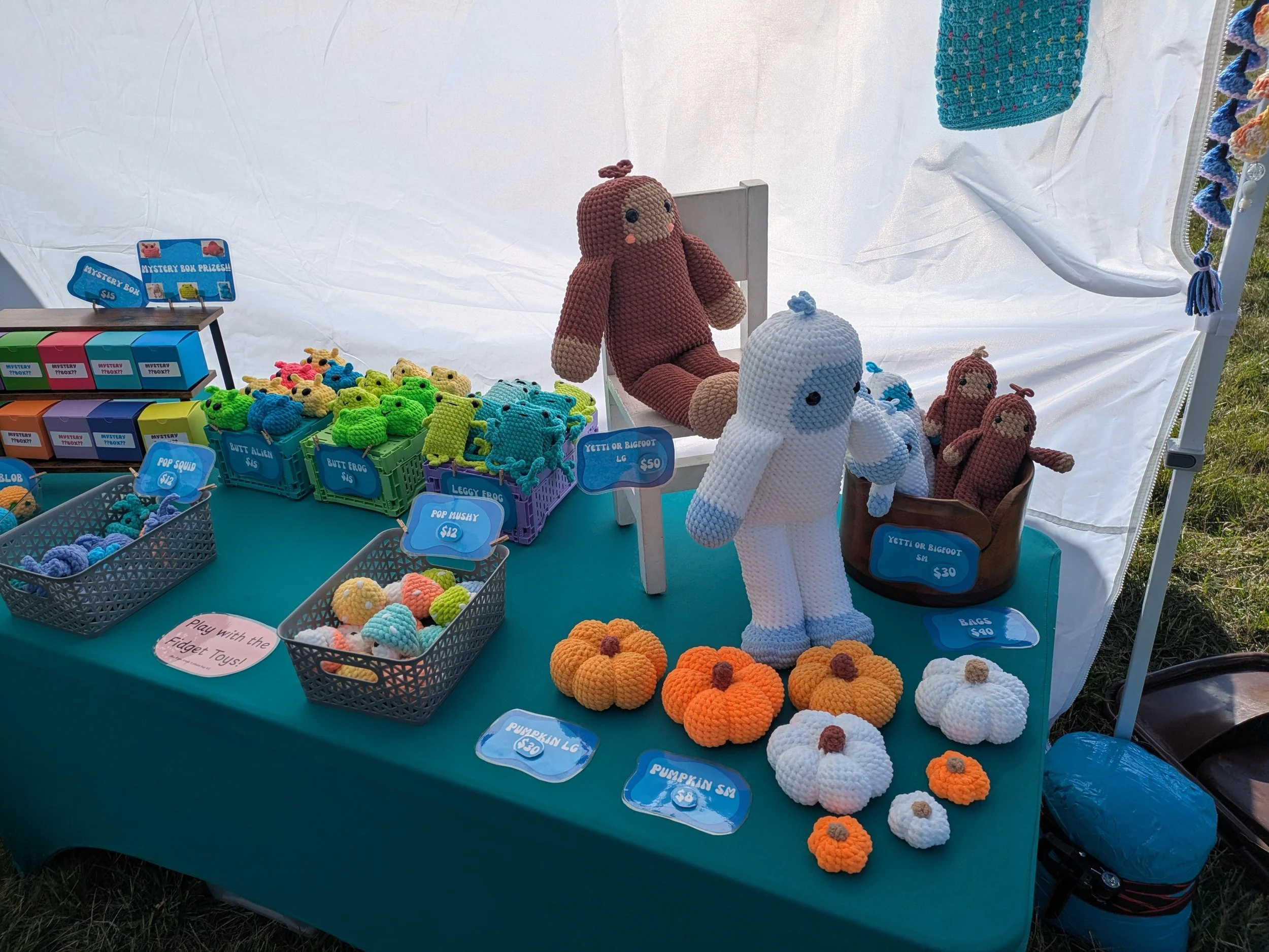 Display of crocheted plush toys including pumpkins, monsters, and Yeti or Bigfoot figures at a craft fair stall.