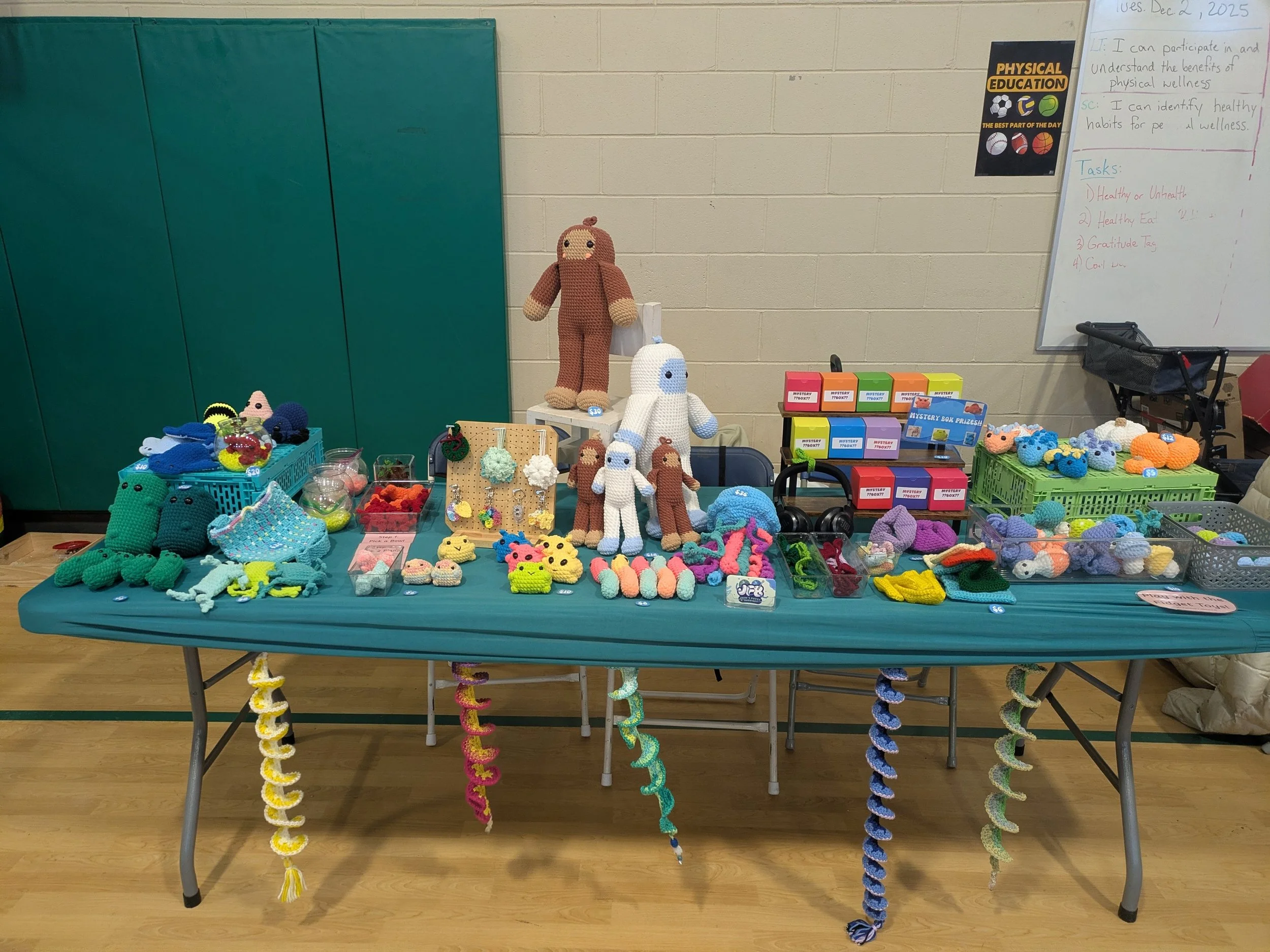 Table display of crocheted toys and accessories at a craft fair, including stuffed animals, keychains, and baskets of colorful handmade items, with large crochet figures standing behind the table and a whiteboard with notes on physical education in the background.