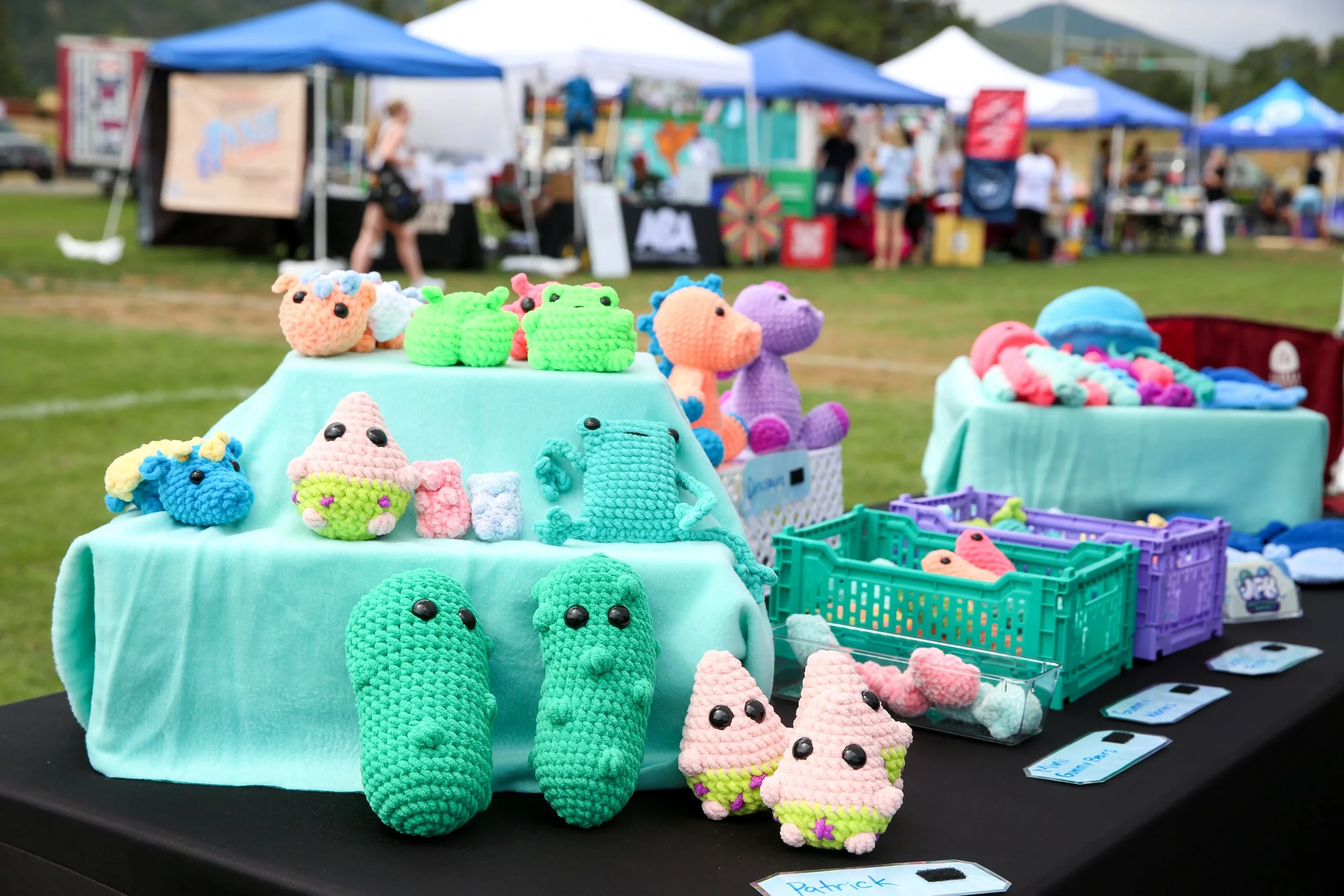 Colorful crochet toys, including pickles, Patrick star, animals, and other plush items, displayed on tables at an outdoor market or festival with tents and people in the background.