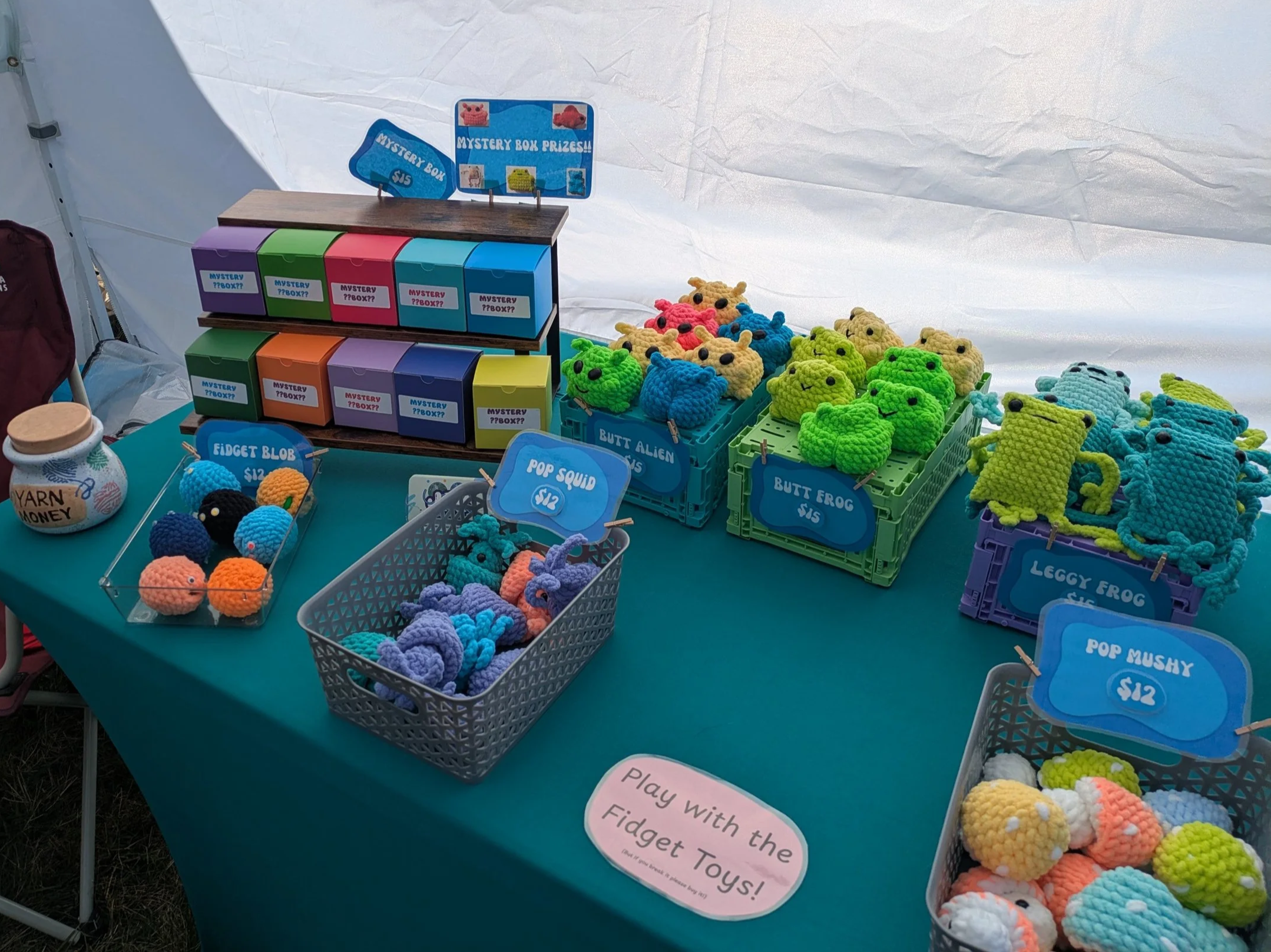 A table at a fair stall displaying crocheted plush toys shaped like aliens, frogs, and squids with labels and price signs, along with colorful mystery boxes and yarn jars.