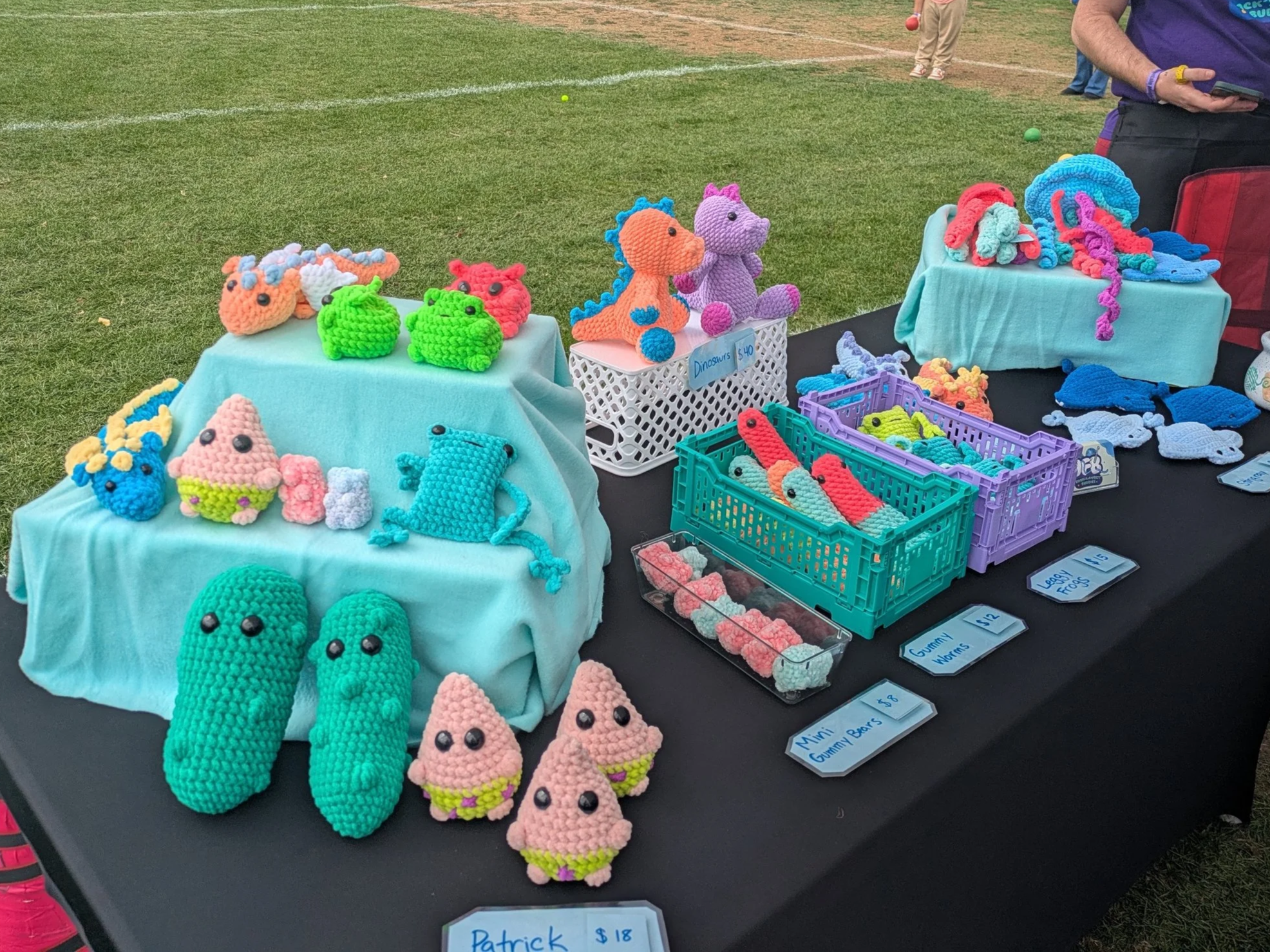 Display of colorful crocheted stuffed animals, leggy frogs, dinosaurs, dragons, jelly fish, frogs and aliens with butts, pickles  at an outdoor market stall, with small baskets and price tags.