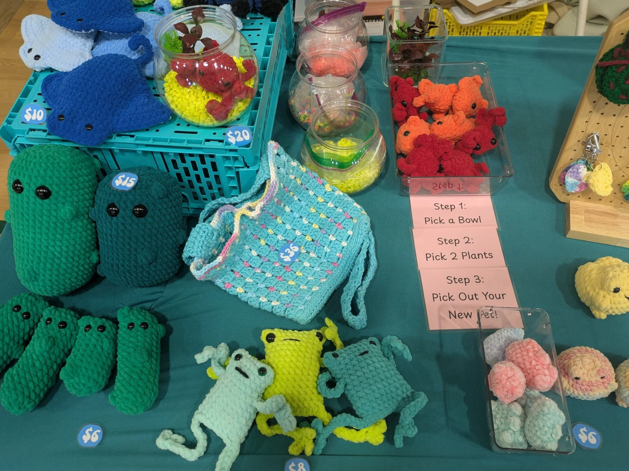 A display of colorful crocheted toys and accessories, including plush animals, small baskets, and plant pots, with a sign outlining steps to pick a bowl, two plants, and a pet.