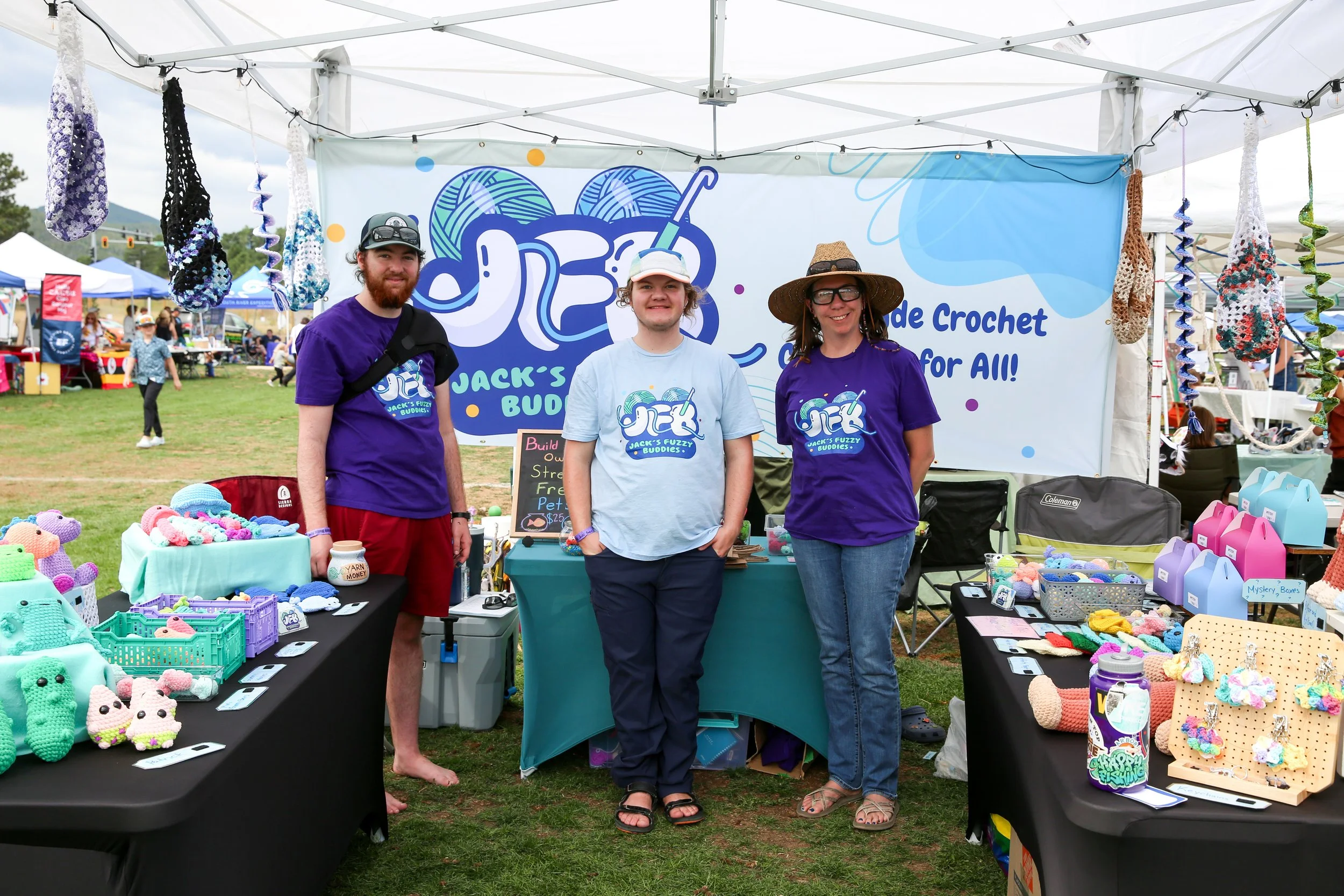 Three people standing in front of a crochet craft booth at an outdoor market. Jacks partner on left jack in middle and friend on the Right