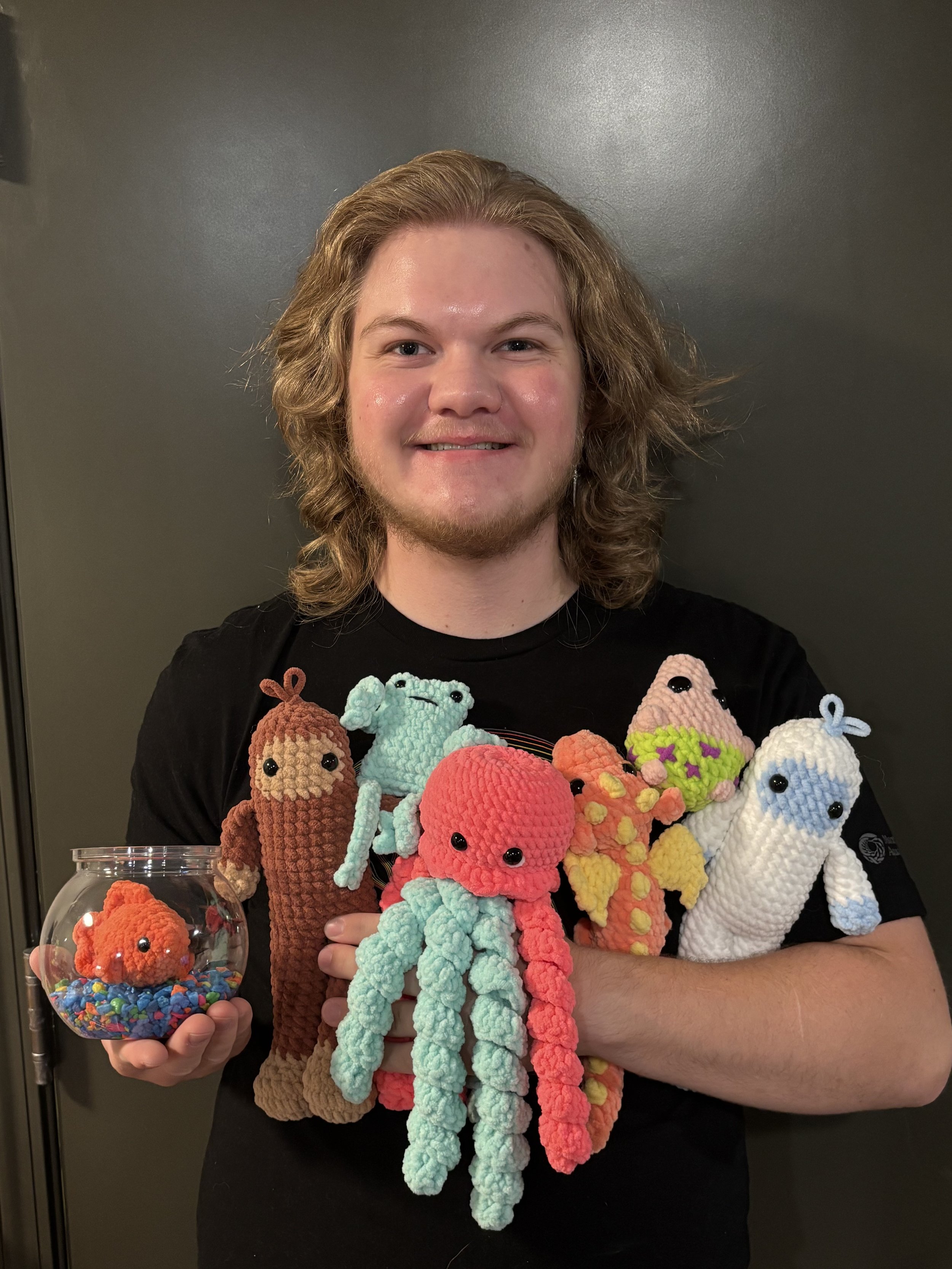 A young man (jack) holding various colorful crocheted octopus toys and a fishbowl with a crocheted octopus inside.