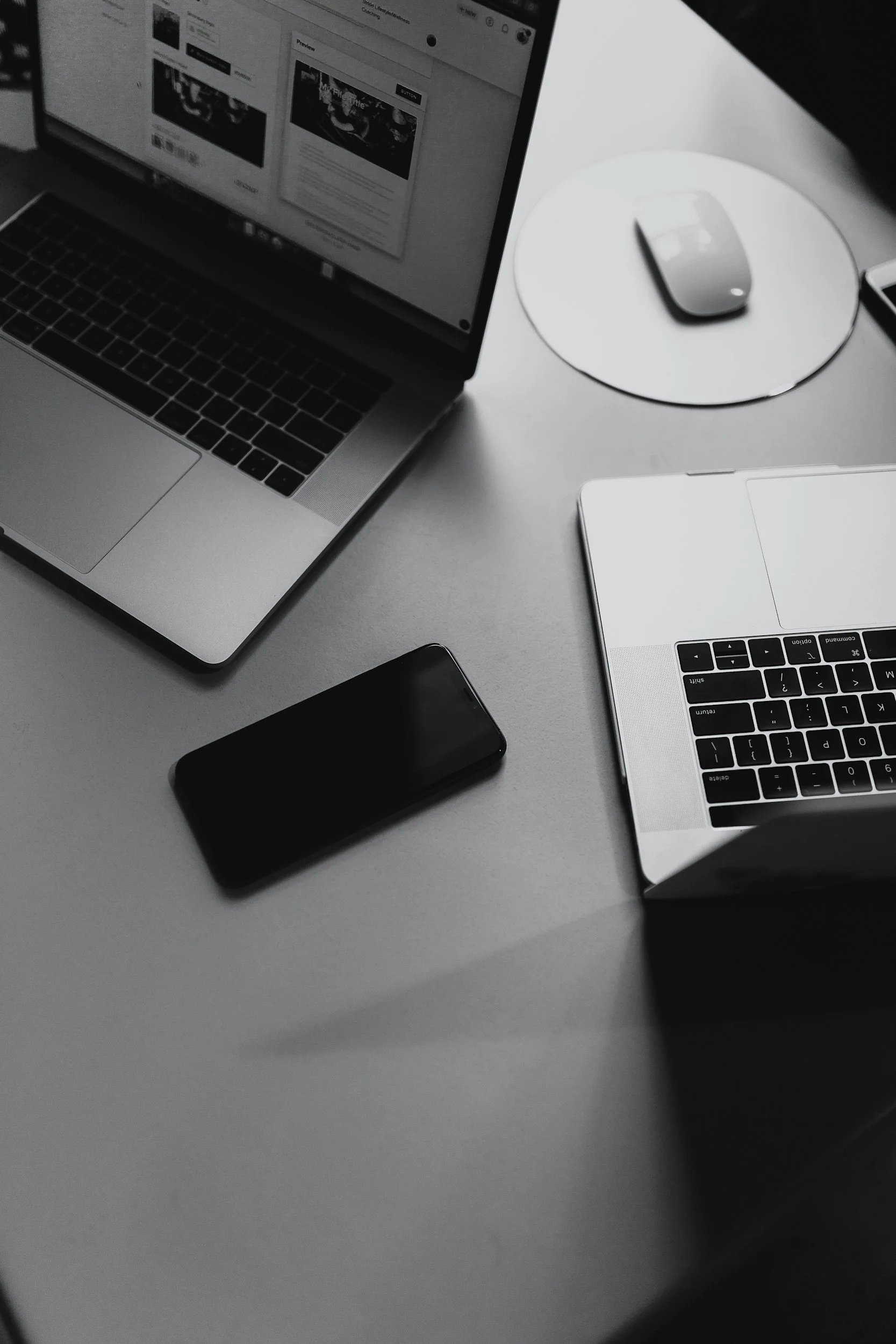 A desk with two open laptops, a smartphone, a wireless mouse on a round mouse pad, and a closed tablet in black and white.