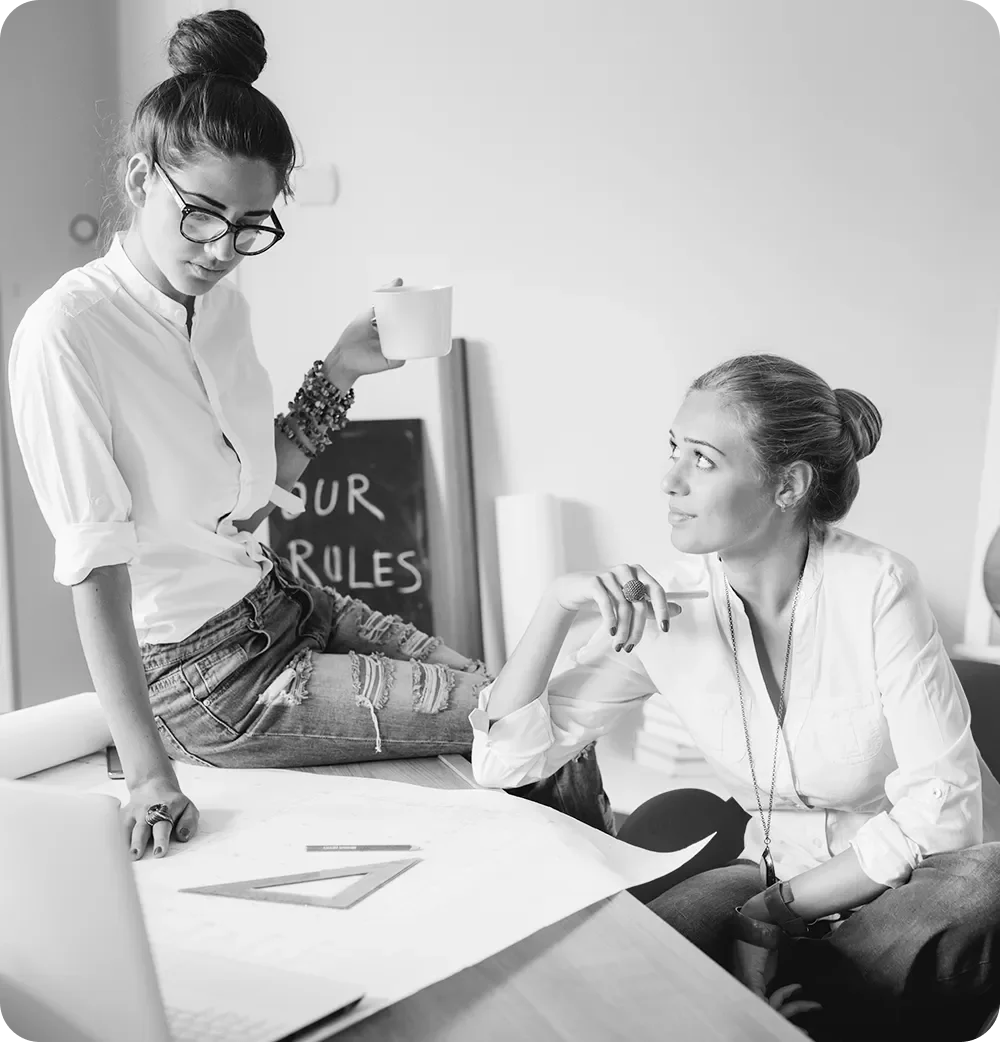 Two women having a conversation in a modern office, with one sitting on a table and the other standing, holding a coffee mug. They appear engaged in a discussion, with a blackboard that says 'OUR RULES' in the background.