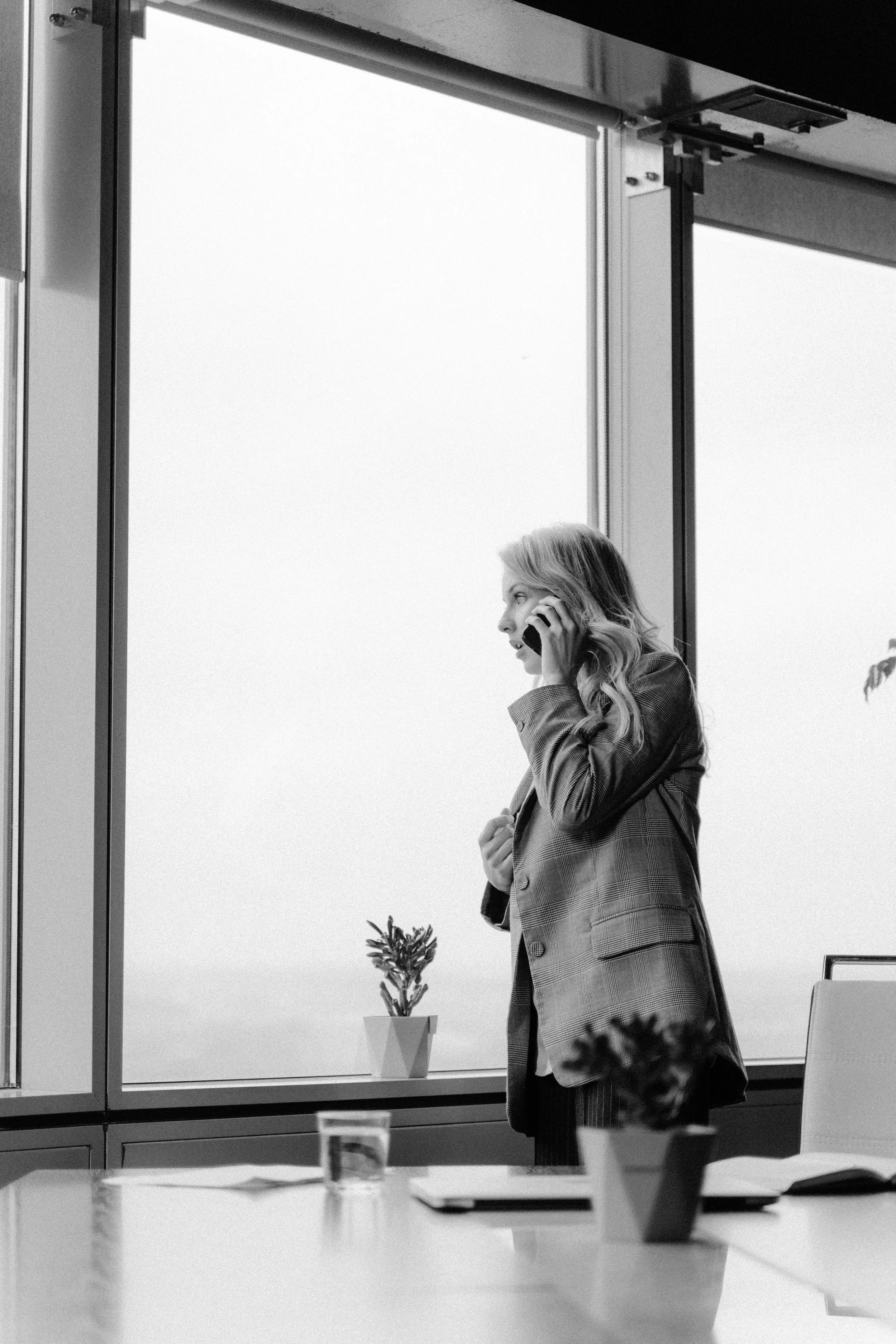 A woman with long hair talking on a cellphone in an office, standing near a large window with a small potted plant on the windowsill, in black and white.