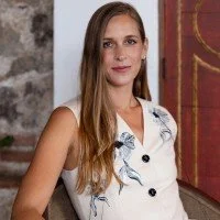 Young woman with long hair wearing a white sleeveless top with embroidery details, standing indoors against a stone wall and a red decorative element.