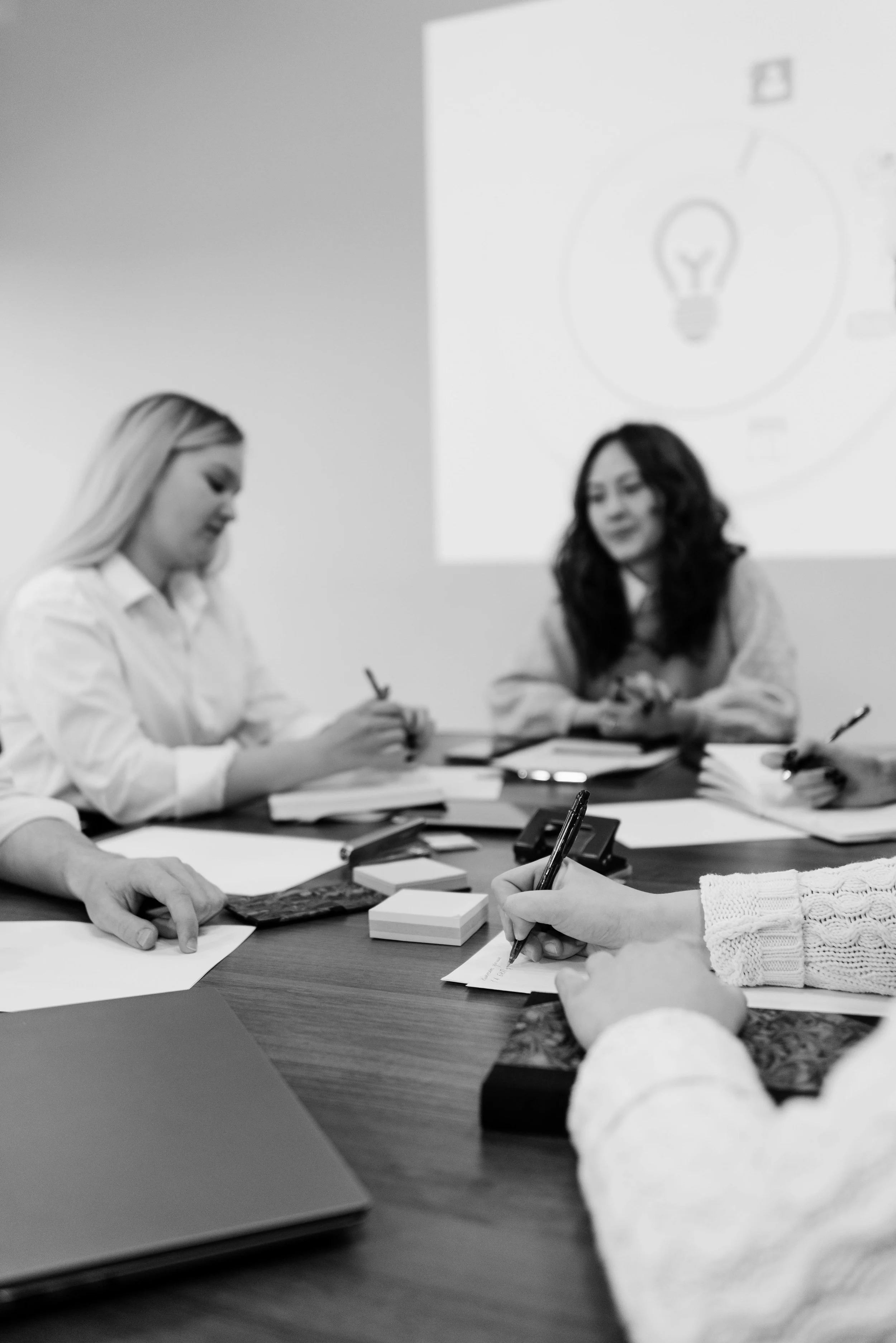 A black and white photo of a business meeting or discussion. Four women are seated around a table with papers, notebooks, and writing instruments. One woman is writing on a notepad, while others are engaged in conversation. There is a large presentation screen in the background showing a light bulb diagram.