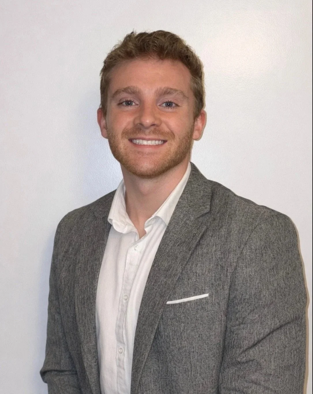 A young man with curly light brown hair and blue eyes, wearing a gray suit jacket and a white shirt, smiling against a plain white background.