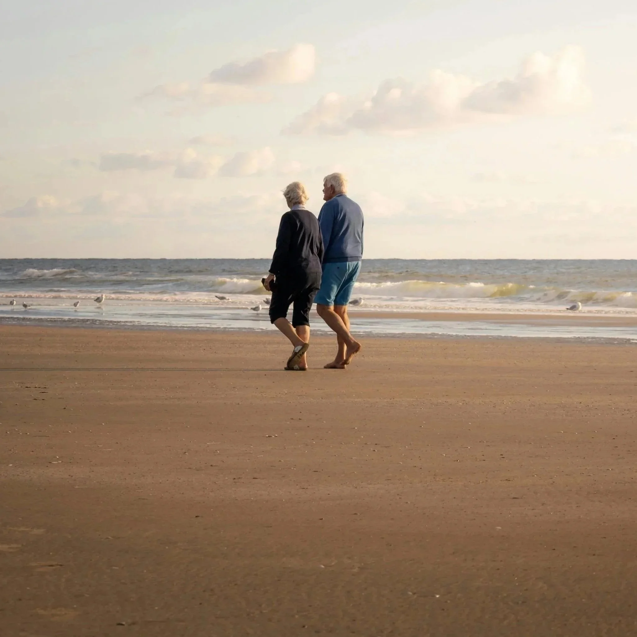 Older couple walking along the beach at sunset, with waves and seagulls in the background.