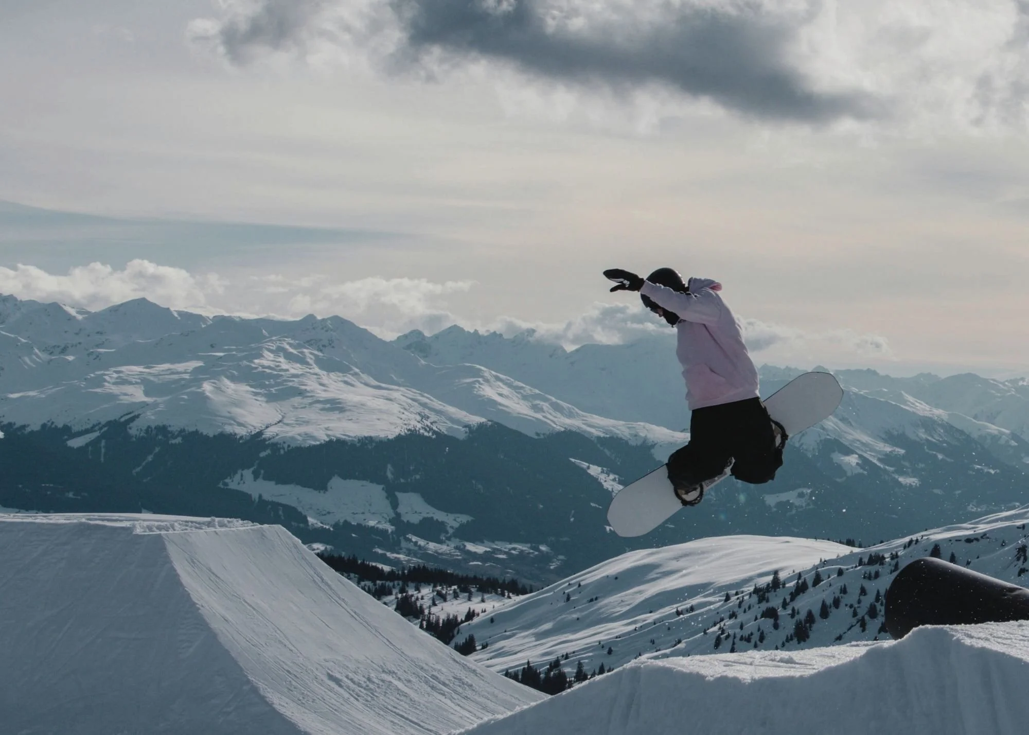 A person snowboarding down a snowy mountain slope in a winter landscape with mountains and cloudy sky in the background.