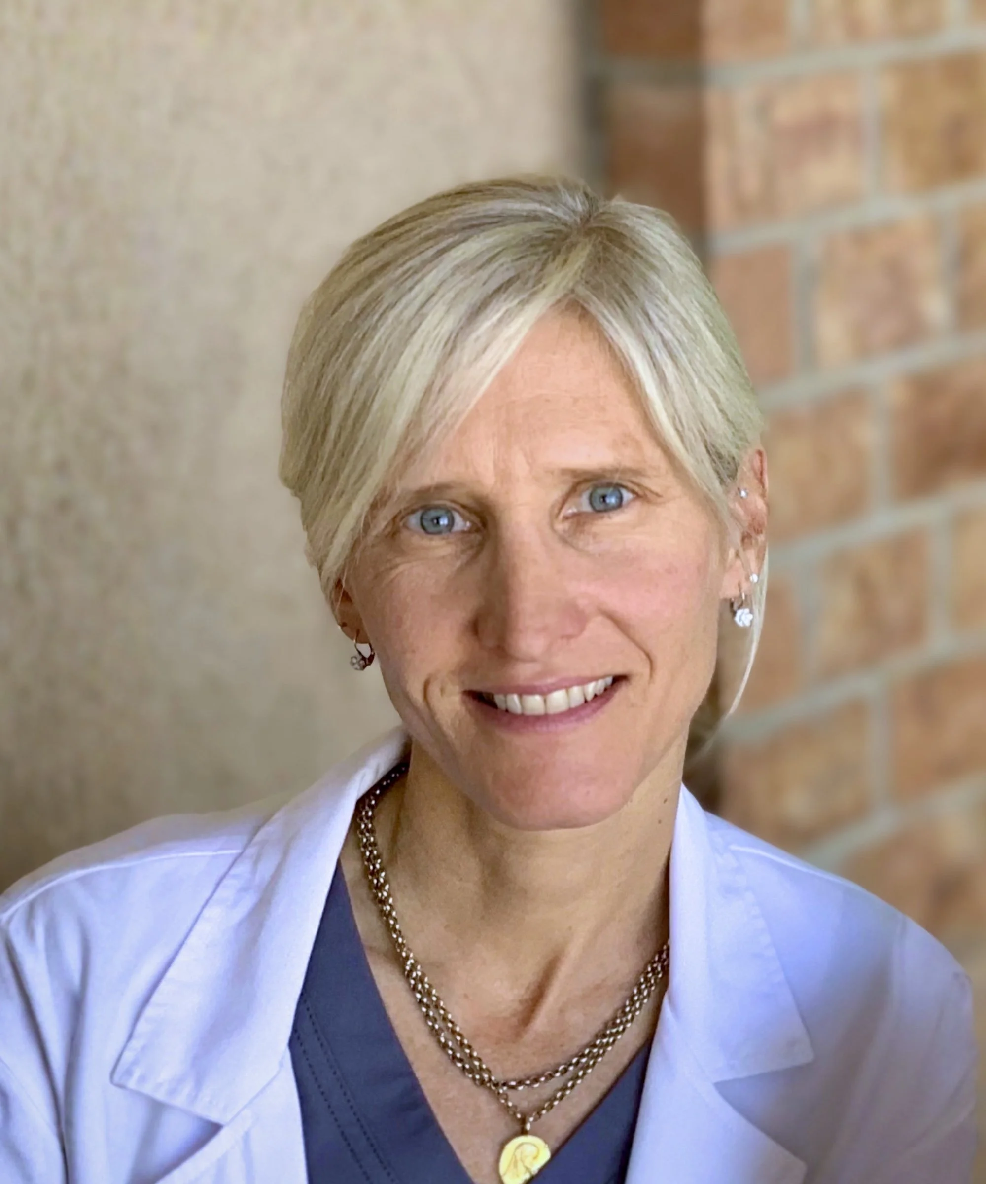 Close-up of a woman with blonde hair, blue eyes, smiling, wearing a white coat, a gray top, and layered gold necklaces with pendants, standing against a brick wall.