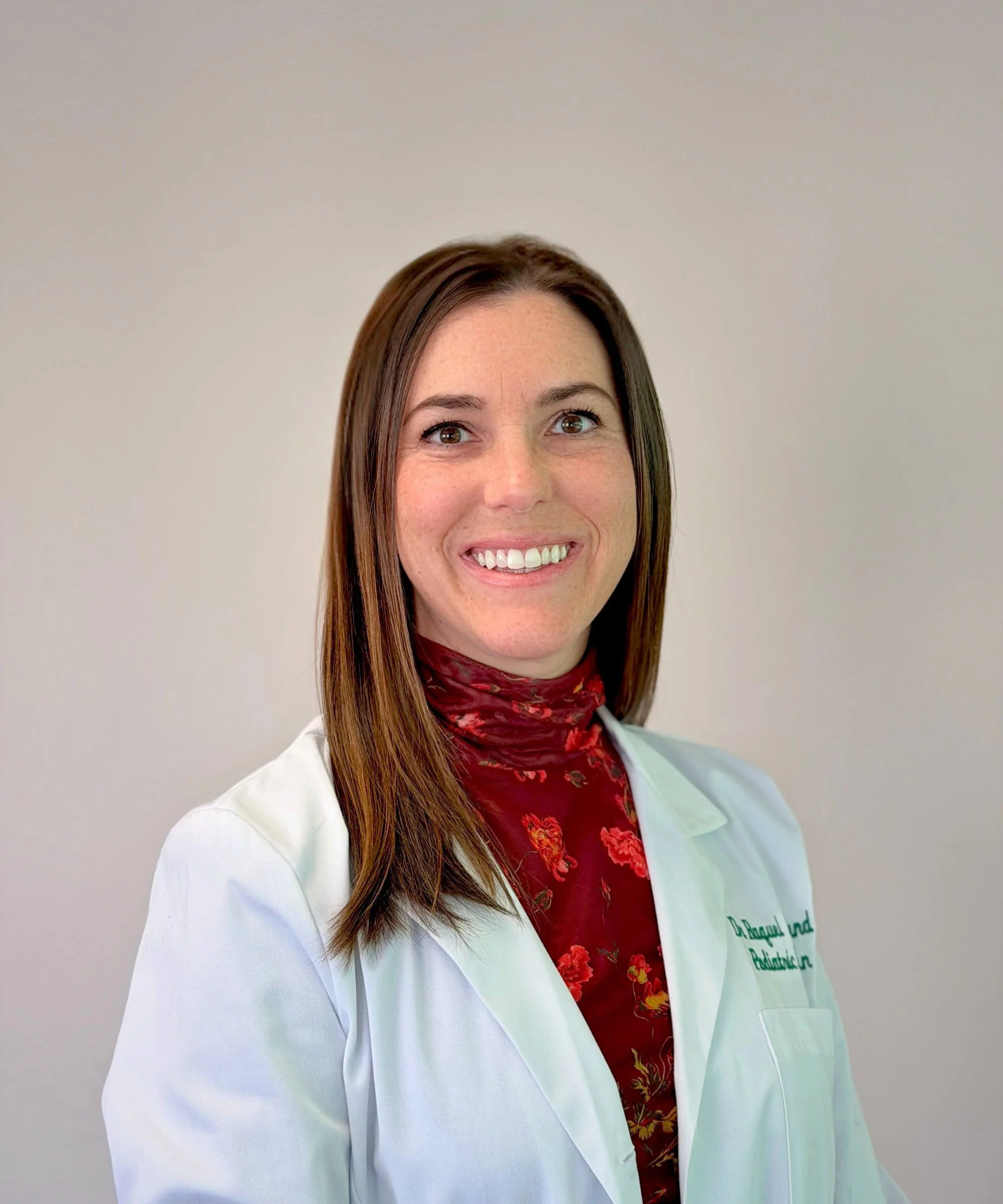 A woman with long brown hair and a bright smile, wearing a white medical coat over a red floral shirt and a red scarf, standing against a plain light-colored wall.