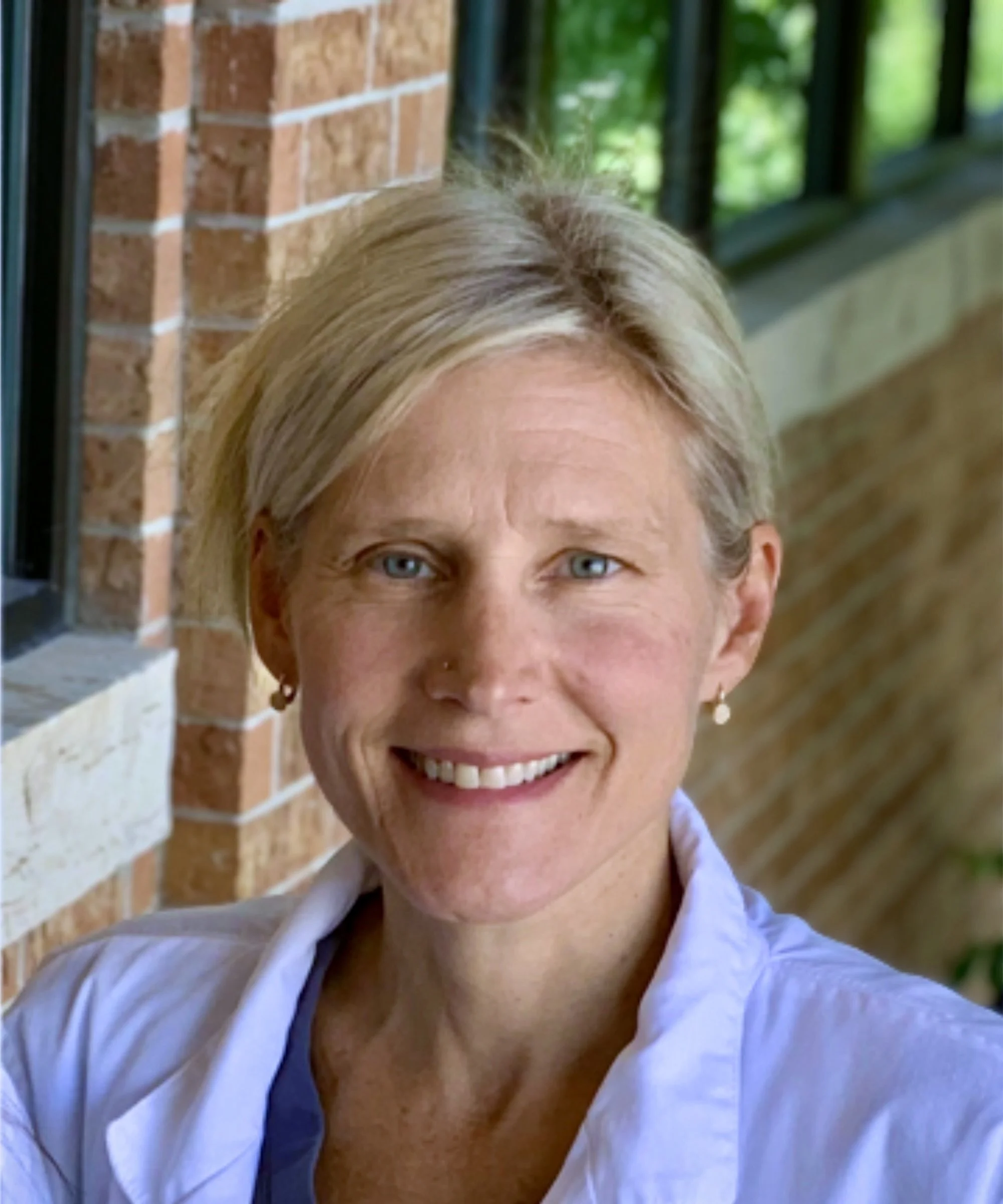Close-up of a smiling woman with blonde hair, blue eyes, wearing a white shirt with a blue strap, standing outside near a brick wall and green foliage.