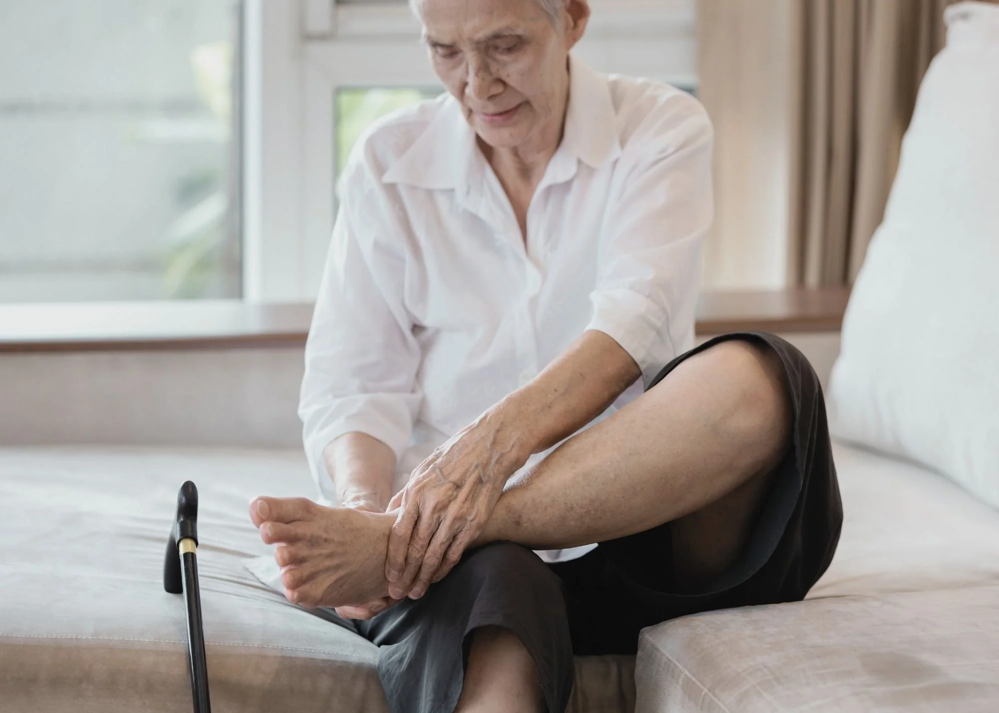 An elderly woman performing physical therapy exercises with a helping hand from a caregiver in a bright room with large windows.