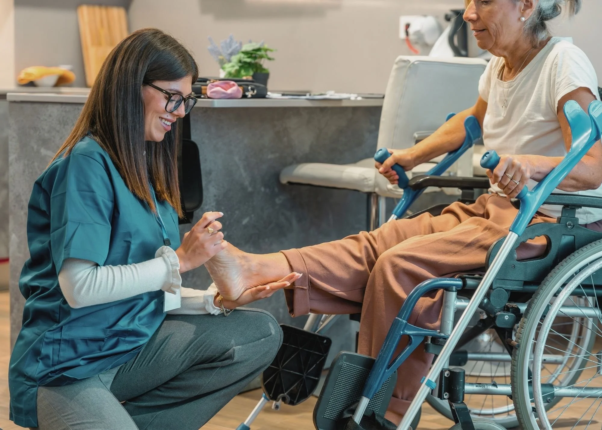 A caregiver kneeling and smiling at an elderly woman in a wheelchair, holding her hands and engaging in a comforting or supportive interaction in a healthcare or nursing facility.