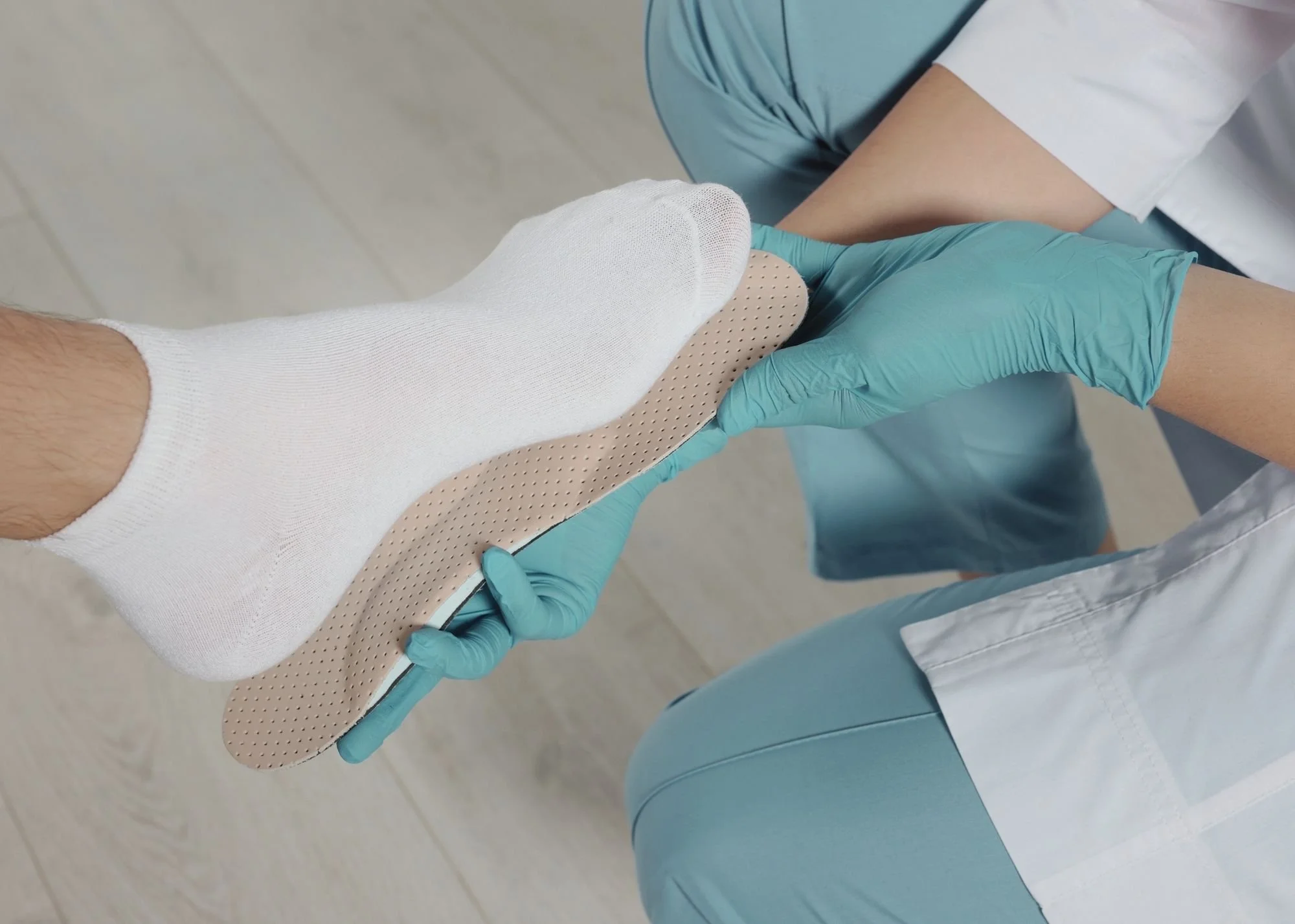 Close-up of a medical professional's gloved hand applying a beige adhesive bandage to a patient's ankle, who is sitting on a blue examination table in a clinical setting.