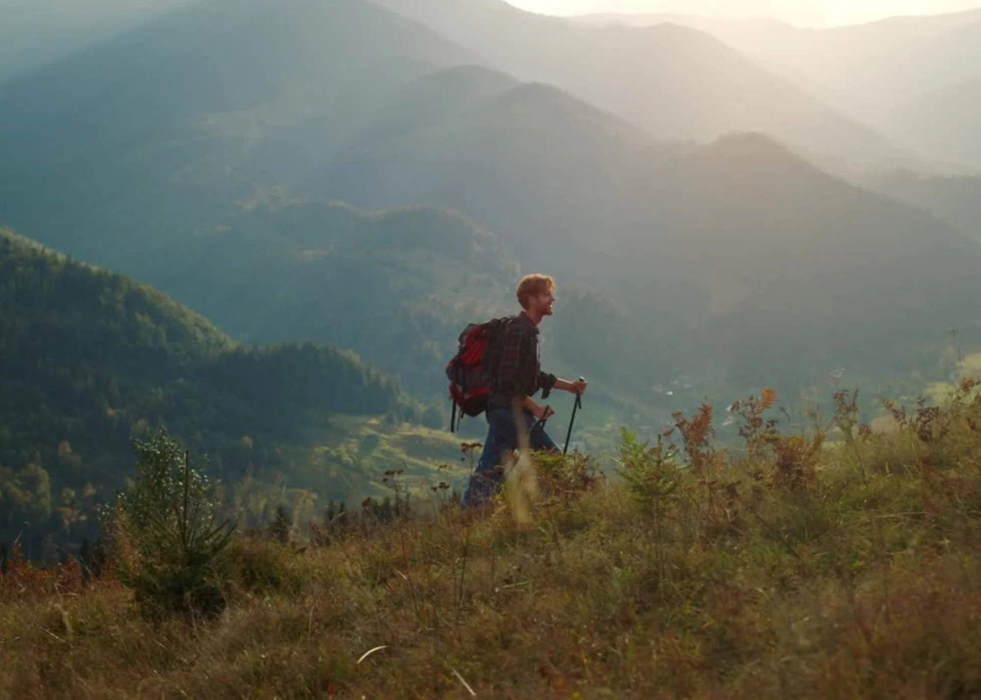 A person with curly hair is hiking in a mountain landscape during sunset, carrying a backpack and walking with trekking poles.