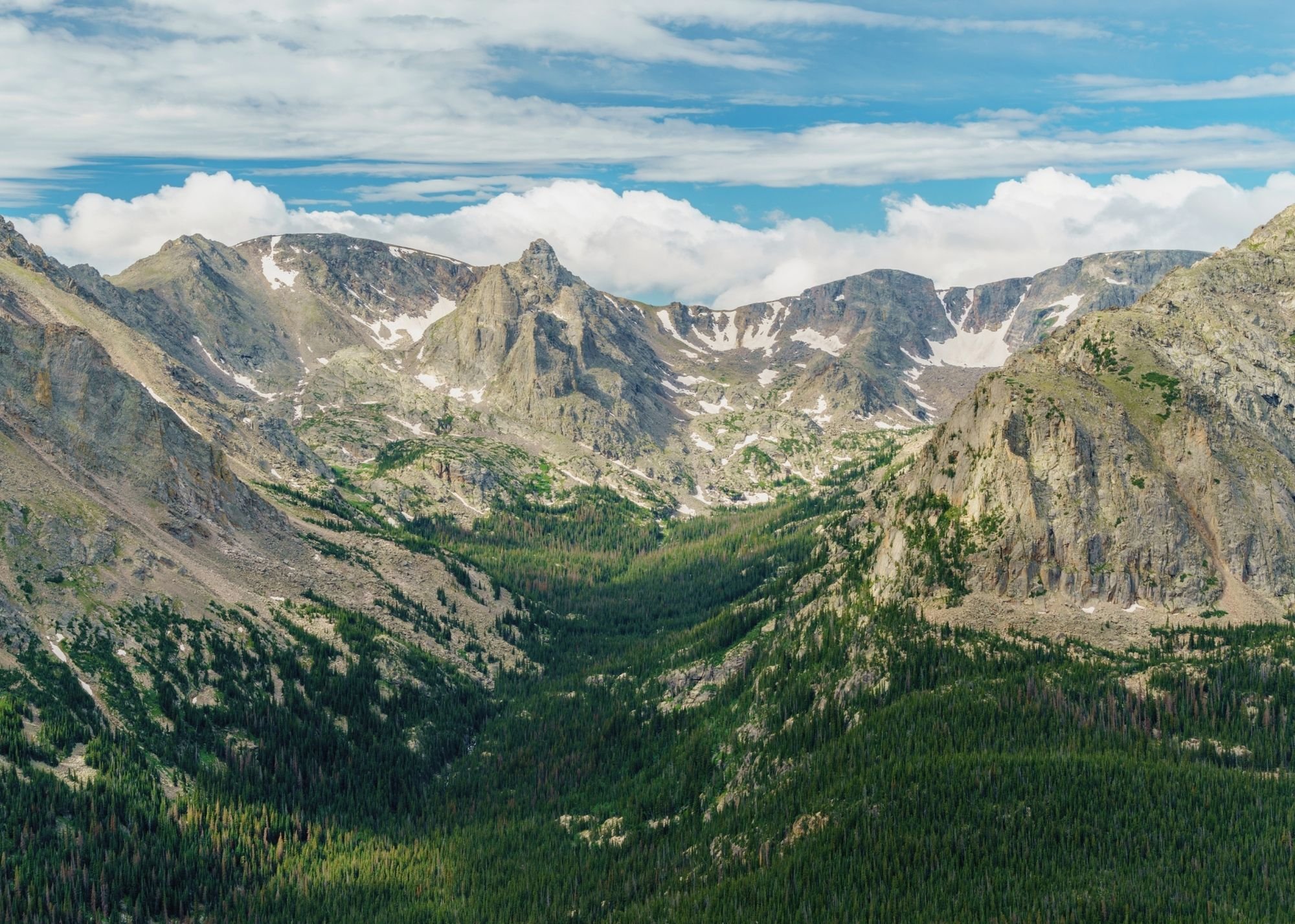 Mountain landscape with rugged peaks, patches of snow, green forested valley, and partly cloudy sky.