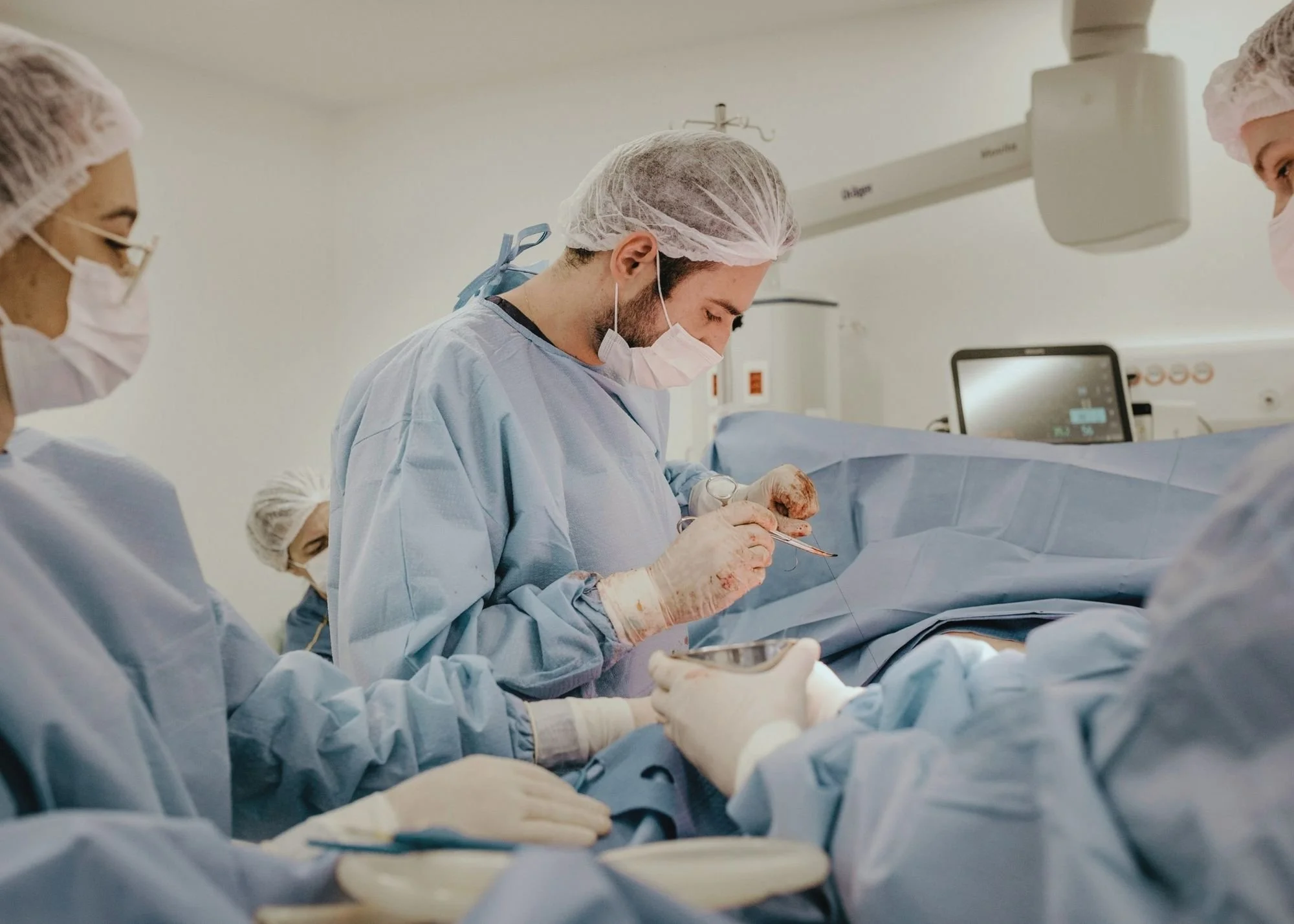 Medical team performing surgery in an operating room, with surgeons dressed in blue scrubs, surgical masks, and hair covers, focused on a procedure.