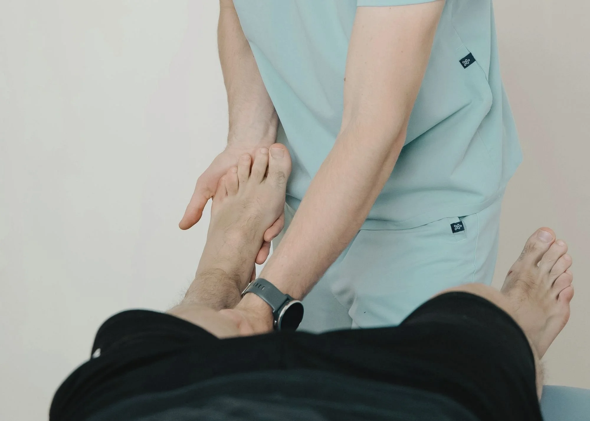 A person in black clothing lying on the ground with their hand extended, being helped up by a person in light blue scrubs, likely a healthcare worker or medical professional.