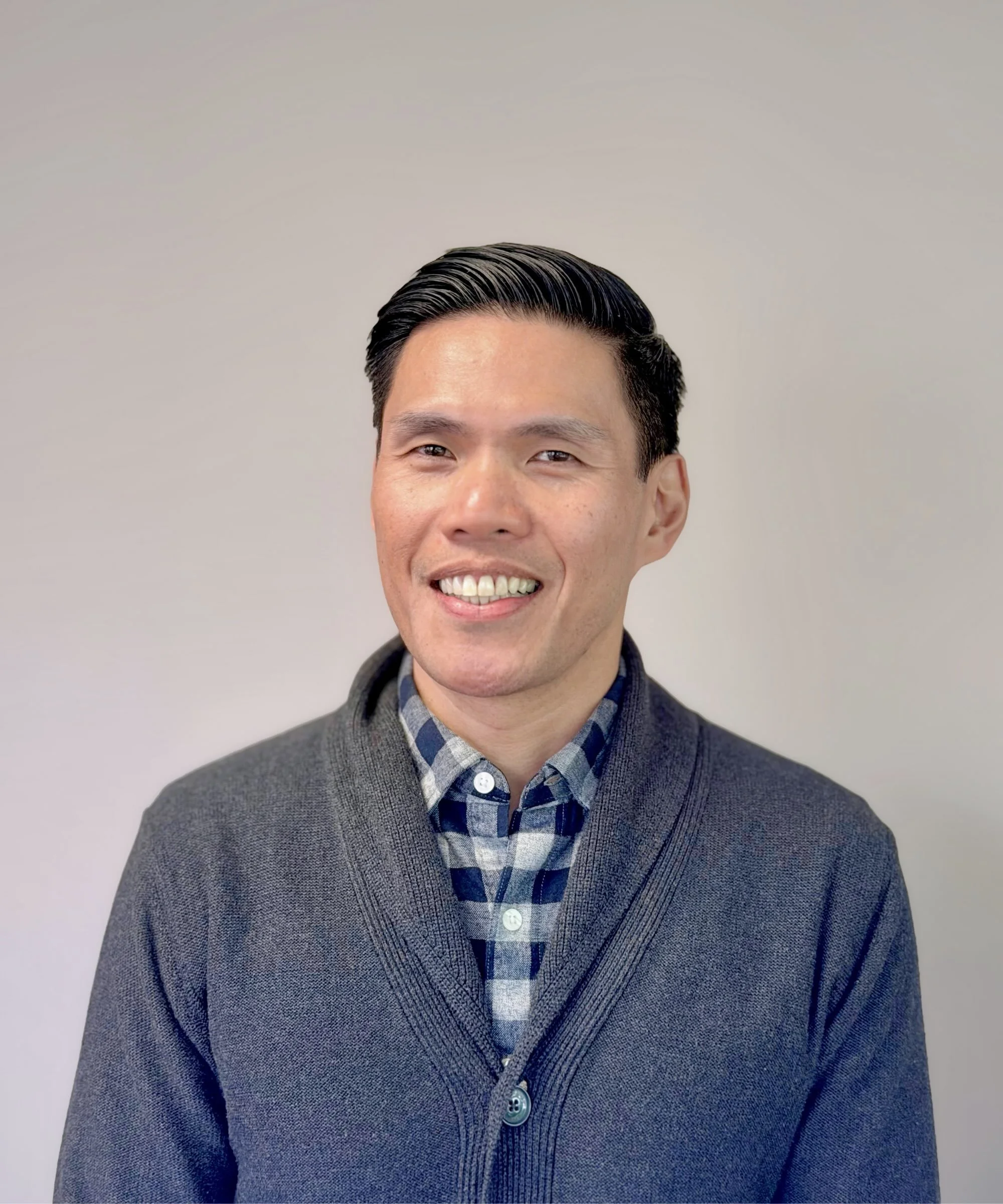 Close-up of a man with black hair and a bright smile, wearing a gray cardigan over a blue plaid shirt, standing against a plain off-white background.