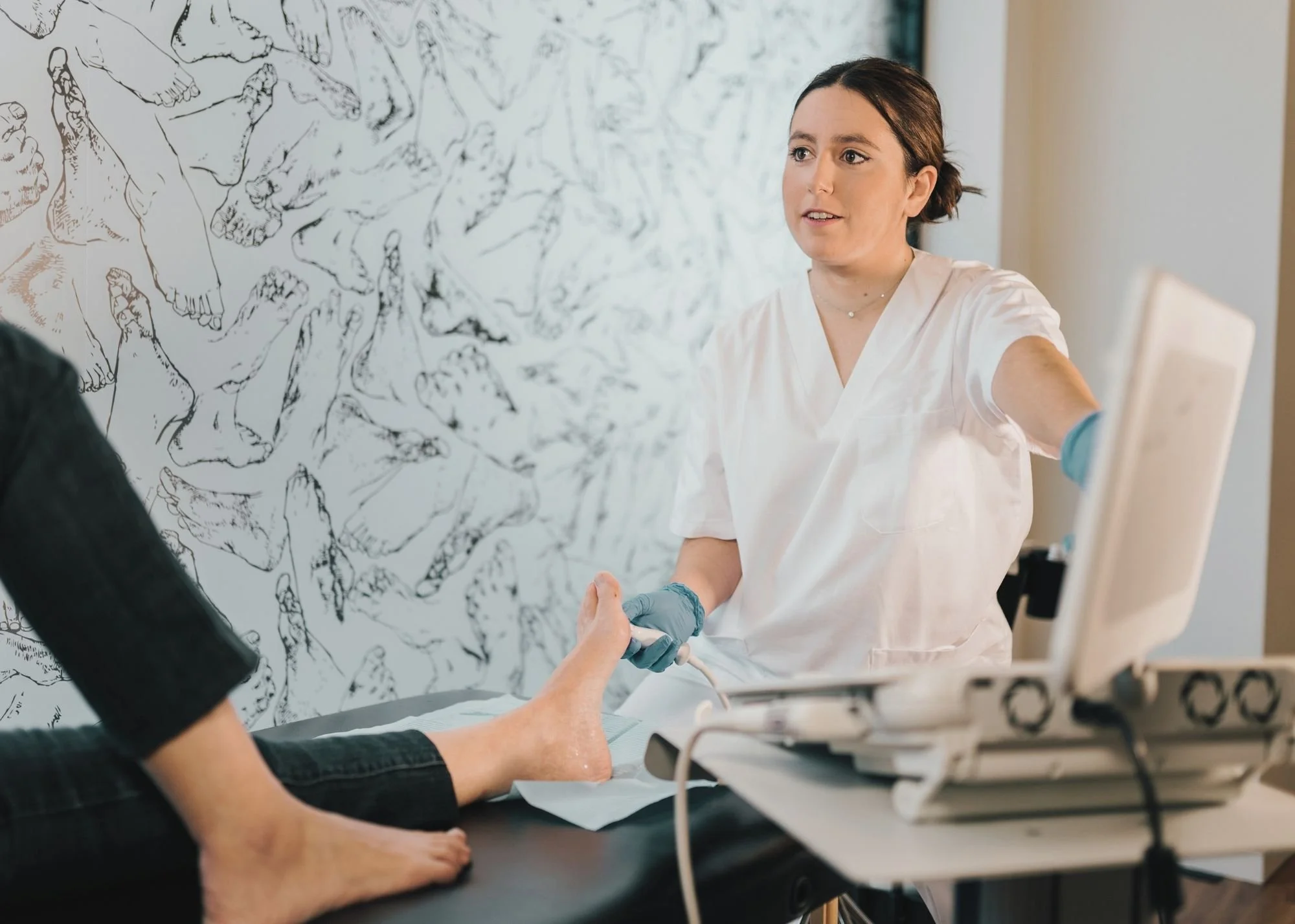 A woman in healthcare scrubs and gloves receiving an IV injection from a healthcare professional in black clothing, in a medical setting with a wall decorated with artwork.