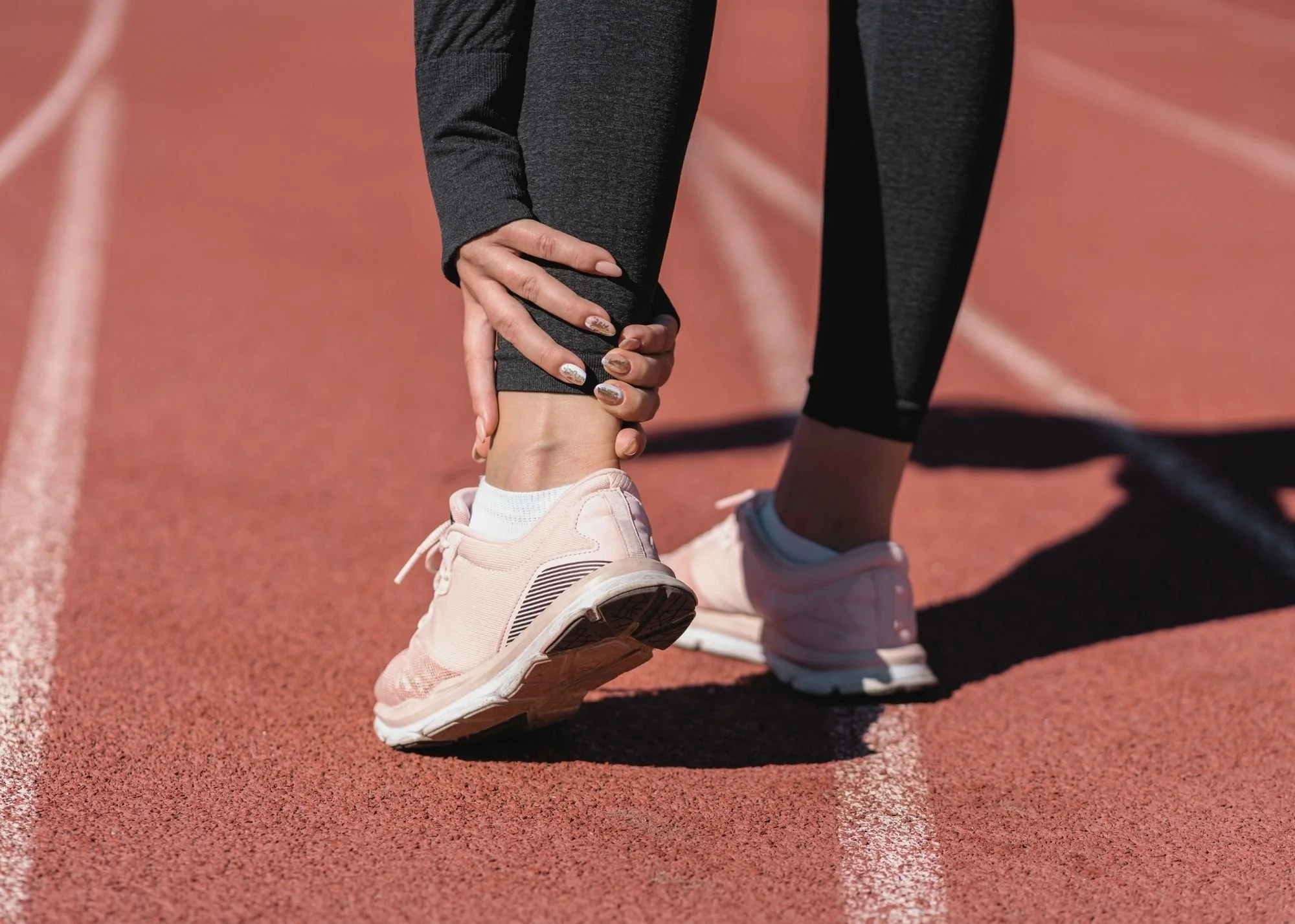 A person in black athletic leggings and pink running shoes standing on a red running track, stretching or adjusting their shoe.