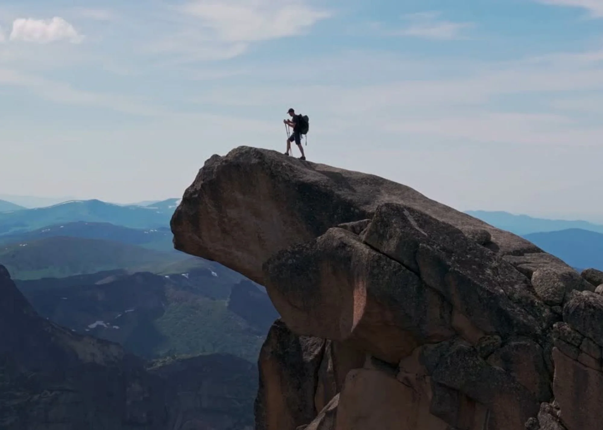 A person standing on a large rock formation in the mountains, surrounded by scenic landscape and blue sky with clouds.