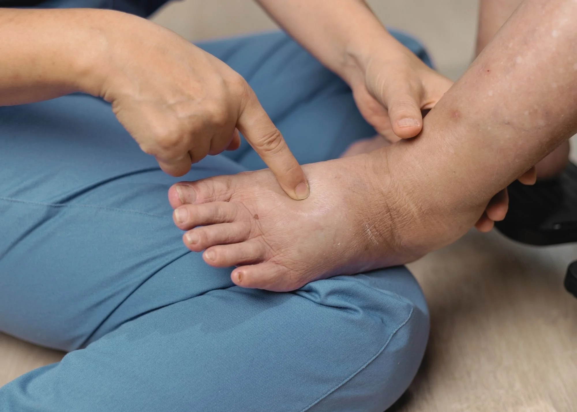 Close-up of a person pointing to their ankle while sitting on the floor, wearing blue pants.