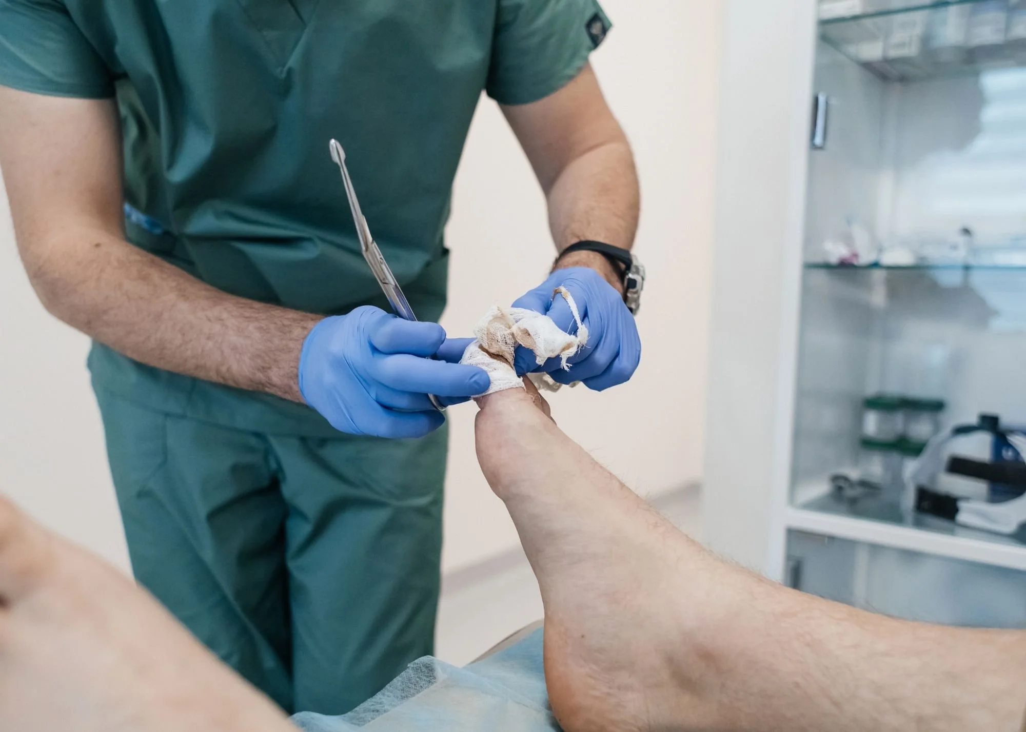 A healthcare professional wearing green scrubs and blue gloves is inserting a medical needle into a patient's arm during a clinical procedure in a medical setting.