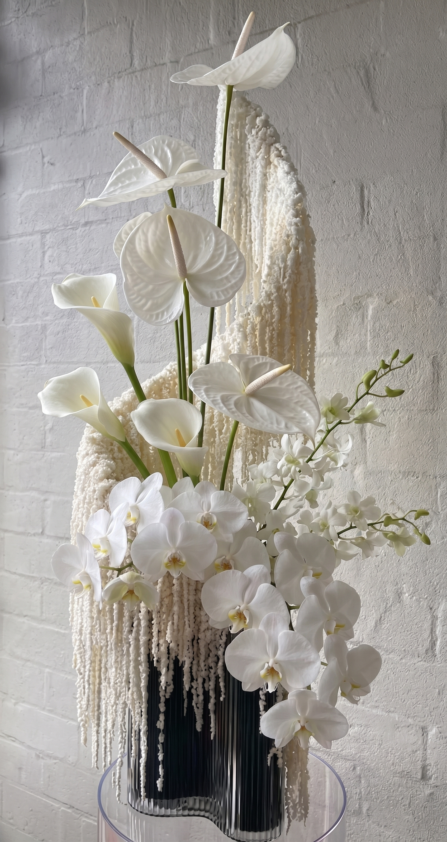 White floral arrangement featuring anthuriums, calla lilies, orchids, and decorative hanging white elements in a black vase against a white brick wall.