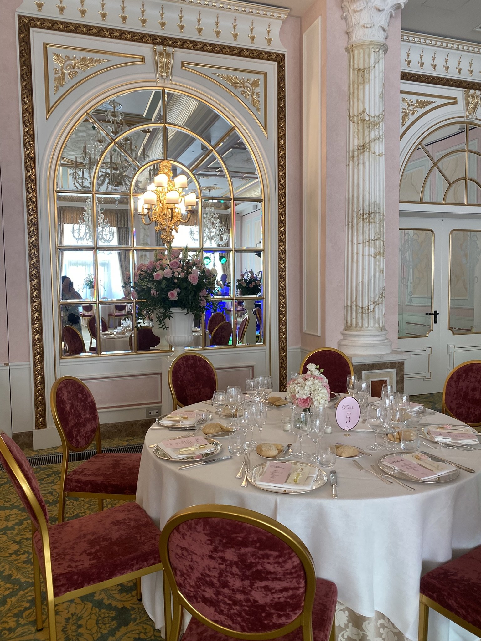 Elegant banquet hall with a round table set for a meal, featuring pink floral centerpieces, gold accents, ornate gold-framed mirror, chandeliers, and pink velvet chairs with gold trim.