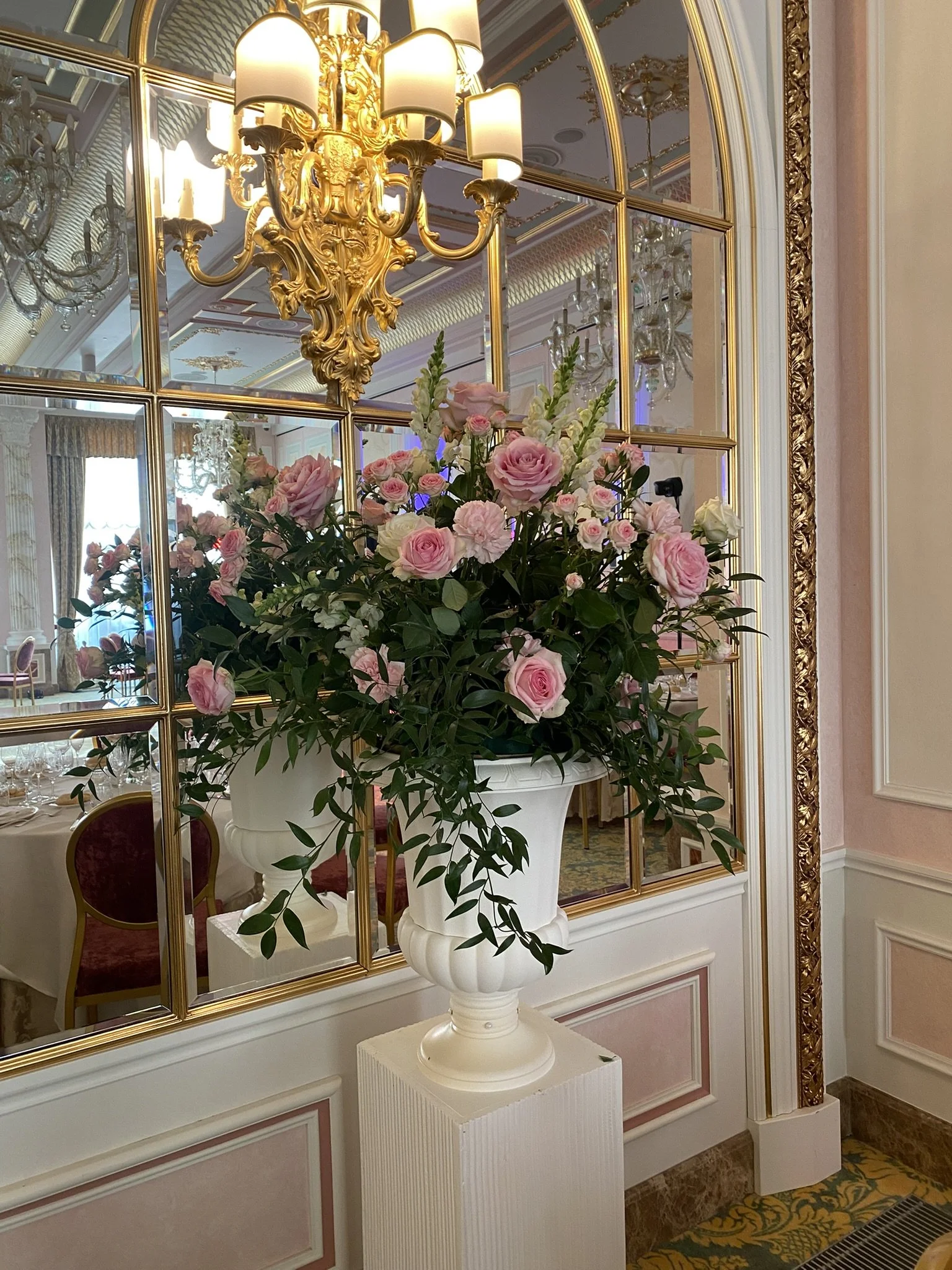 A large floral arrangement of pink roses and greenery in a white pedestal vase, set against a gold-framed mirror and ornate chandelier reflected in a luxurious, elegant room.