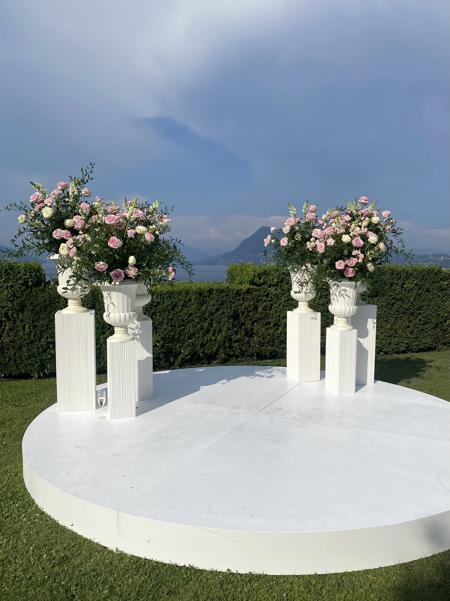 White circular platform with four white pedestals holding flower arrangements of pink and white roses with greenery. In the background, there is greenery, a lake, mountains, and a cloudy sky.