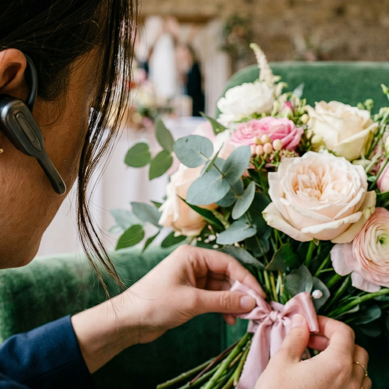 A woman adjusting a pink ribbon on a bouquet of light pink and white roses and green foliage.