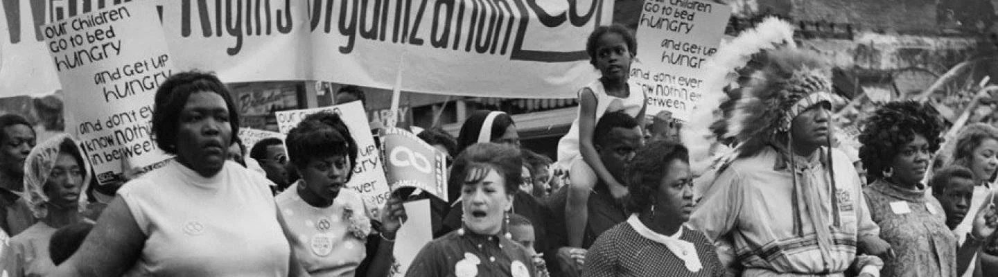 A diverse group of protesters, predominantly women, marching with signs and banners, one of which reads 'Rights Victimize'. One participant wears a Native American headdress. The protest appears to be about social or civil rights issues.