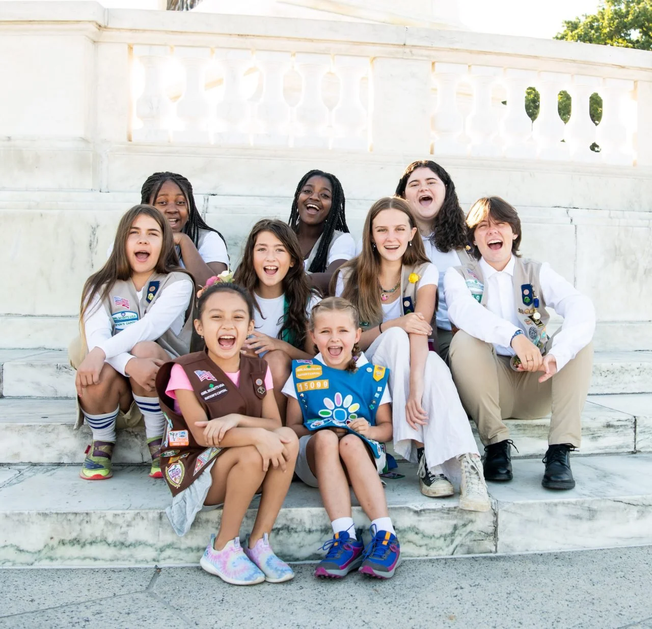 Group of smiling Boy Scouts and Girl Scouts sitting on marble steps outdoors.