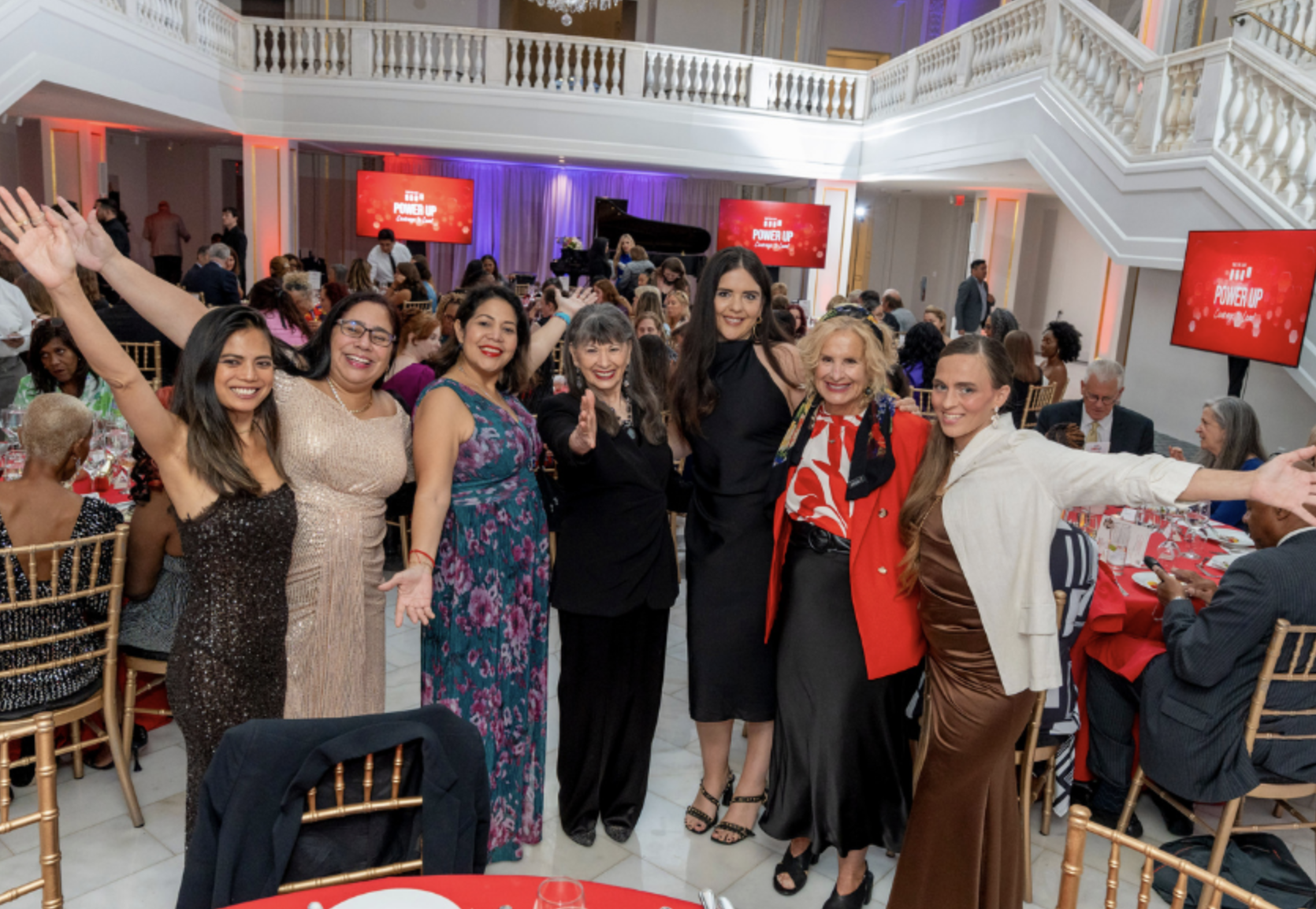 A group of women at a formal event or celebration, standing and smiling with arms raised, in a decorated banquet hall with many seated guests and red tables.